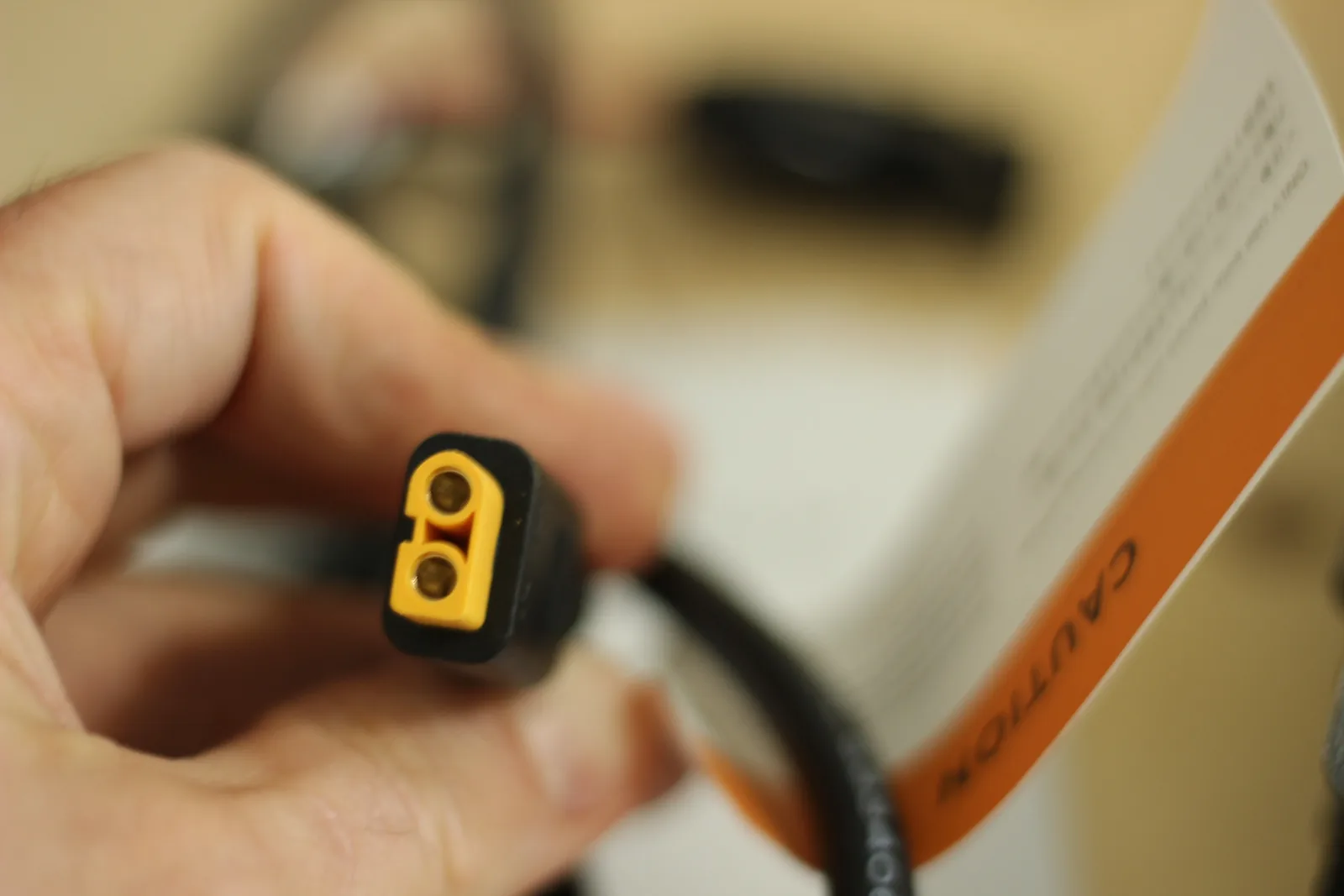 A yellow XT60 connector on a black cable held between finger and thumb, with an orange-banded caution page behind