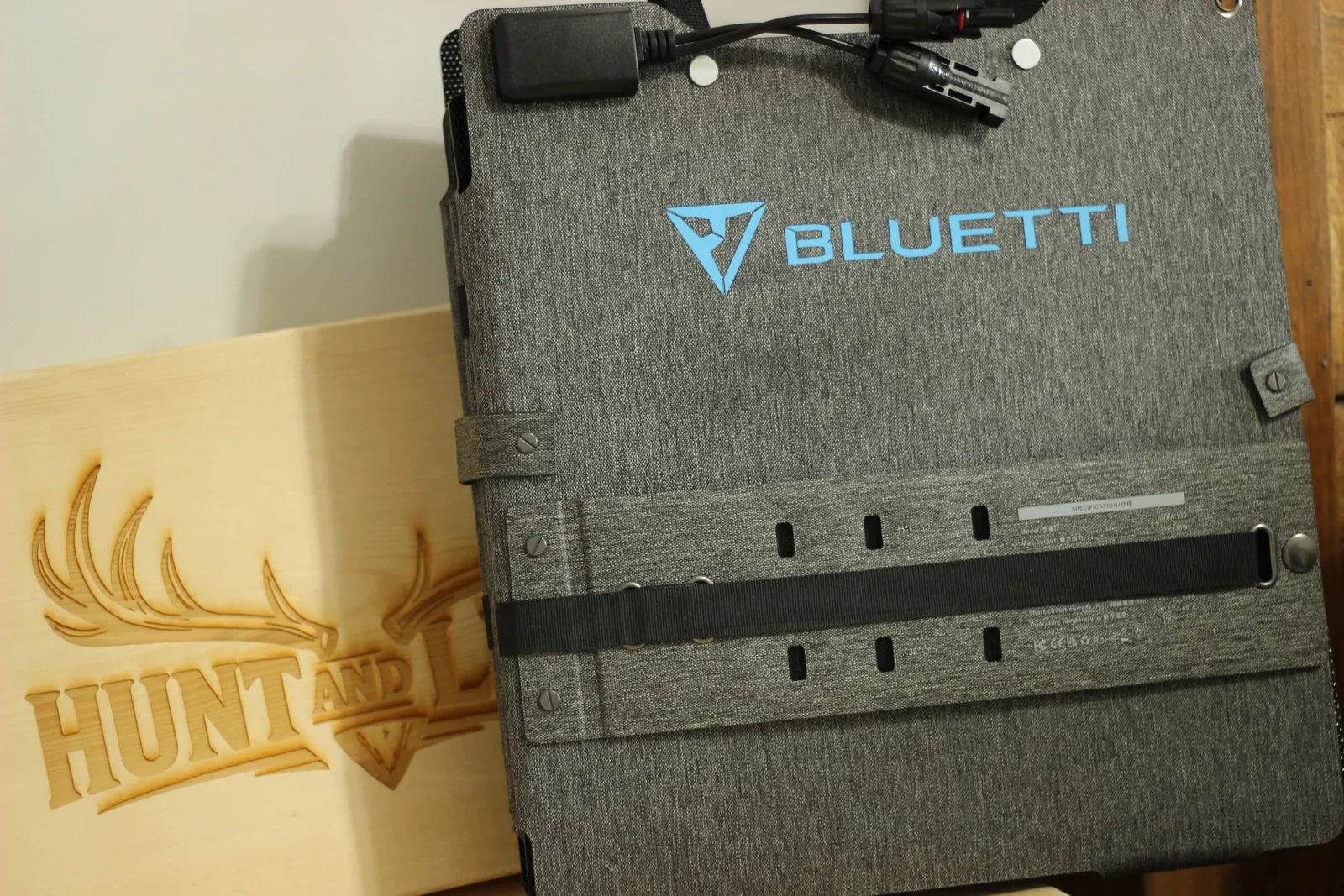 The folded panel standing beside a Hunt and Live engraved wooden plaque, showing the webbing strap, kickstand panels and the printed specification label
