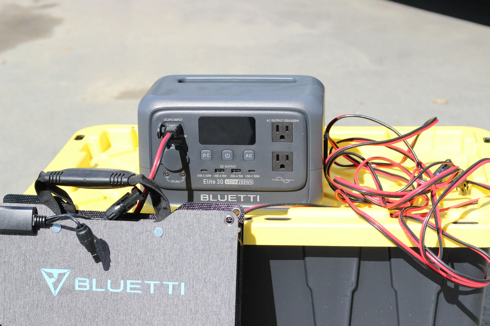 The unfolded solar panel on a driveway with a BLUETTI Elite 30 power station sitting on a yellow tote lid, connected by a red and black lead