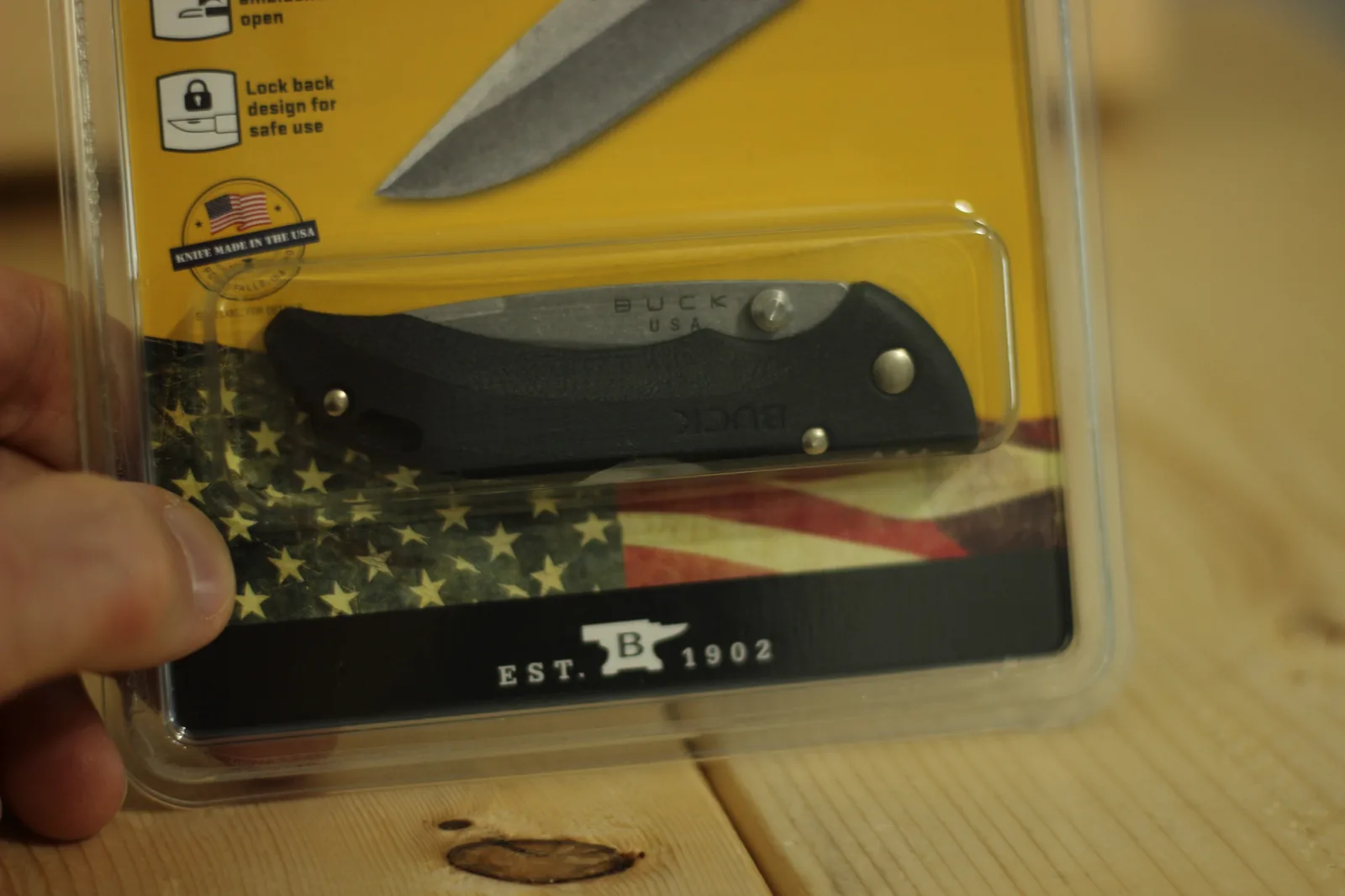 Blister pack held in a hand showing the closed black knife through the plastic, over a printed American flag and a KNIFE MADE IN THE USA seal
