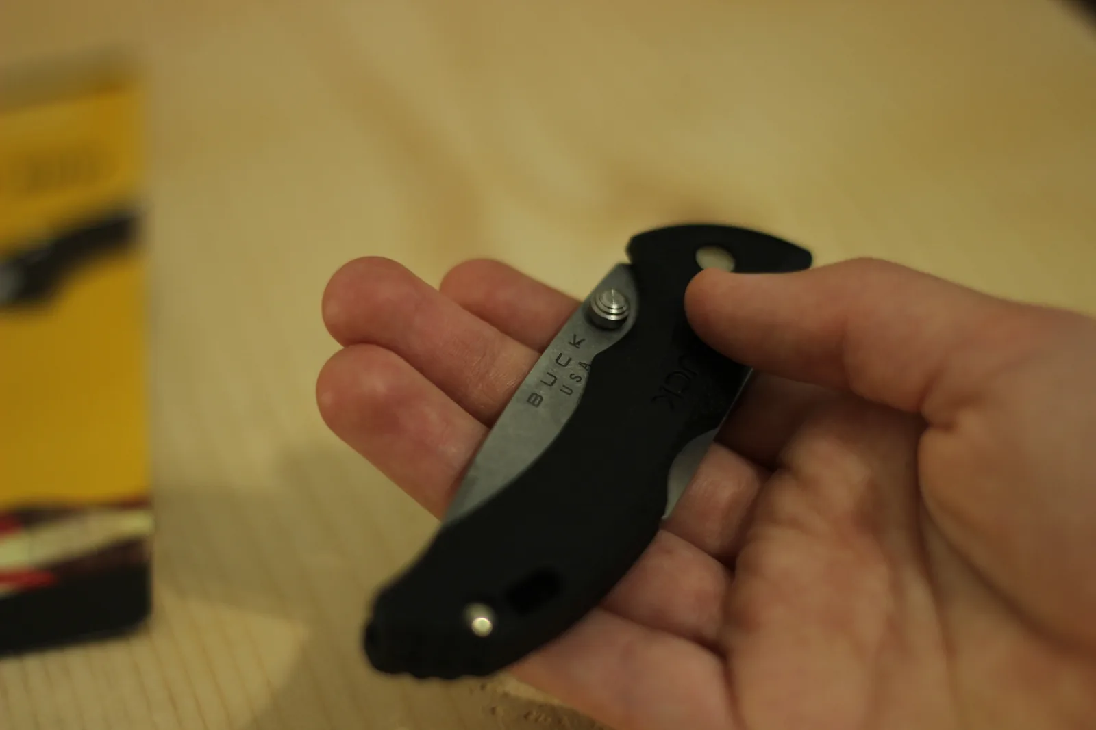 The closed knife held in a hand with the yellow packaging blurred in the background, showing the size relative to the fingers