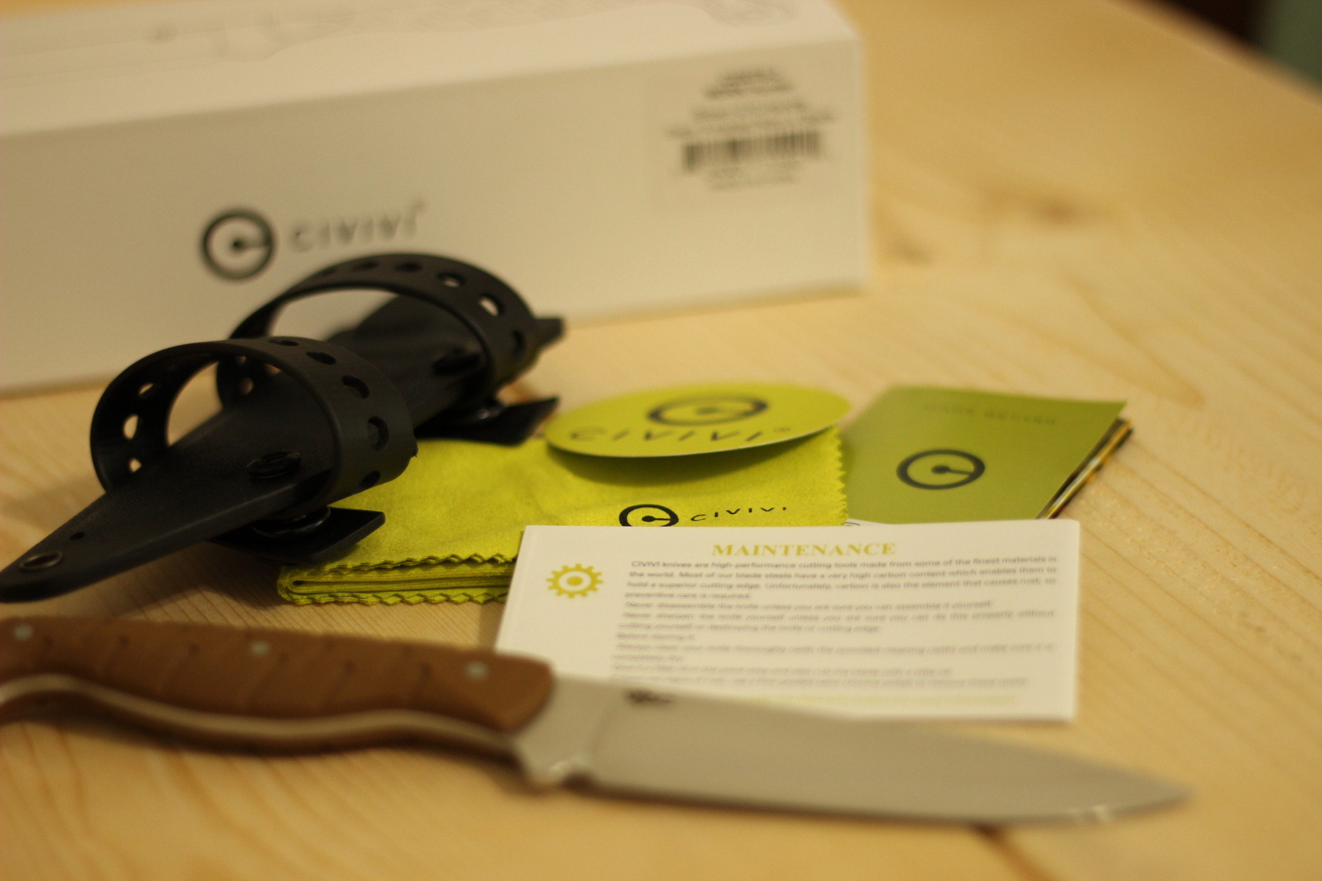 The full unboxing spread on a light wood plank — the kydex sheath with the two polymer carrier straps wrapped around it on the left, a green Civivi microfibre cloth and small Civivi sticker in the centre, a folded warranty leaflet, and the Civivi MDRN Hunter knife laid out at the bottom with its brown G10 handle and bare satin blade exposed