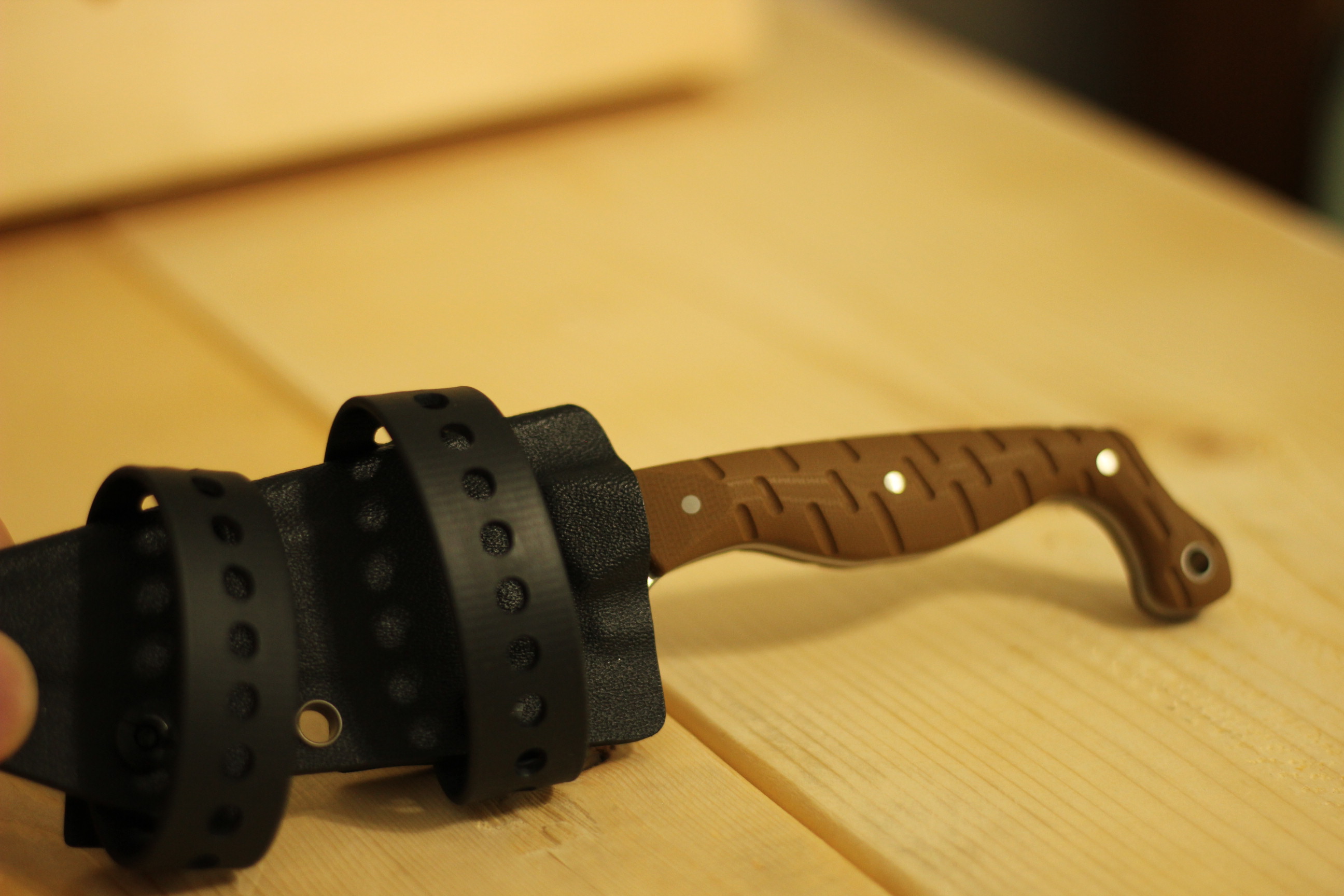 The Civivi MDRN Hunter sheathed in its black kydex sheath with the two polymer carrier straps wrapped around it, laid on a light wood surface with the brown G10 handle visible extending out of the sheath to the right