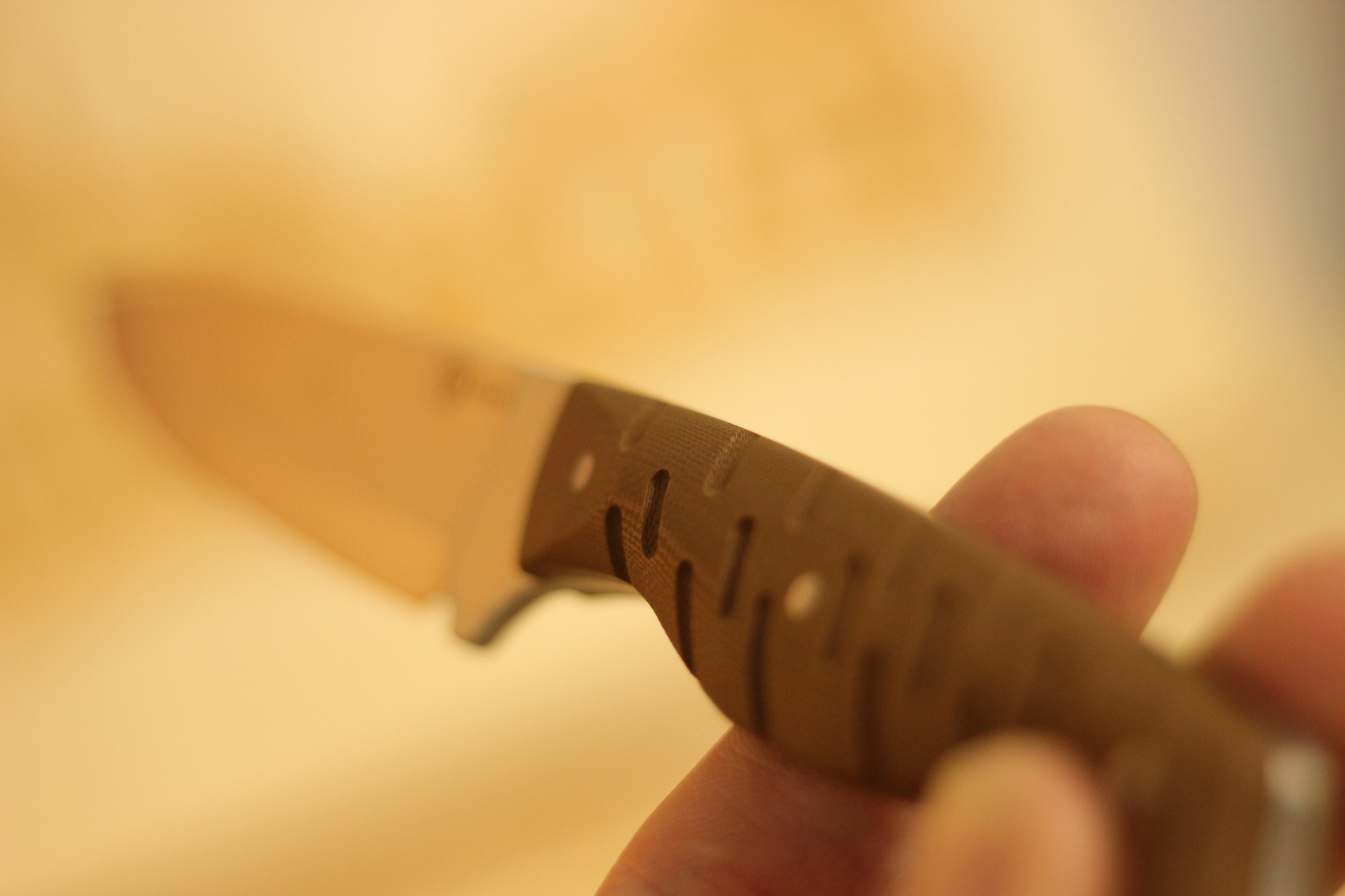 The Civivi MDRN Hunter held in hand against a warm yellow blurred kitchen background — blade and brown G10 handle both in soft focus, showing the overall handling proportion