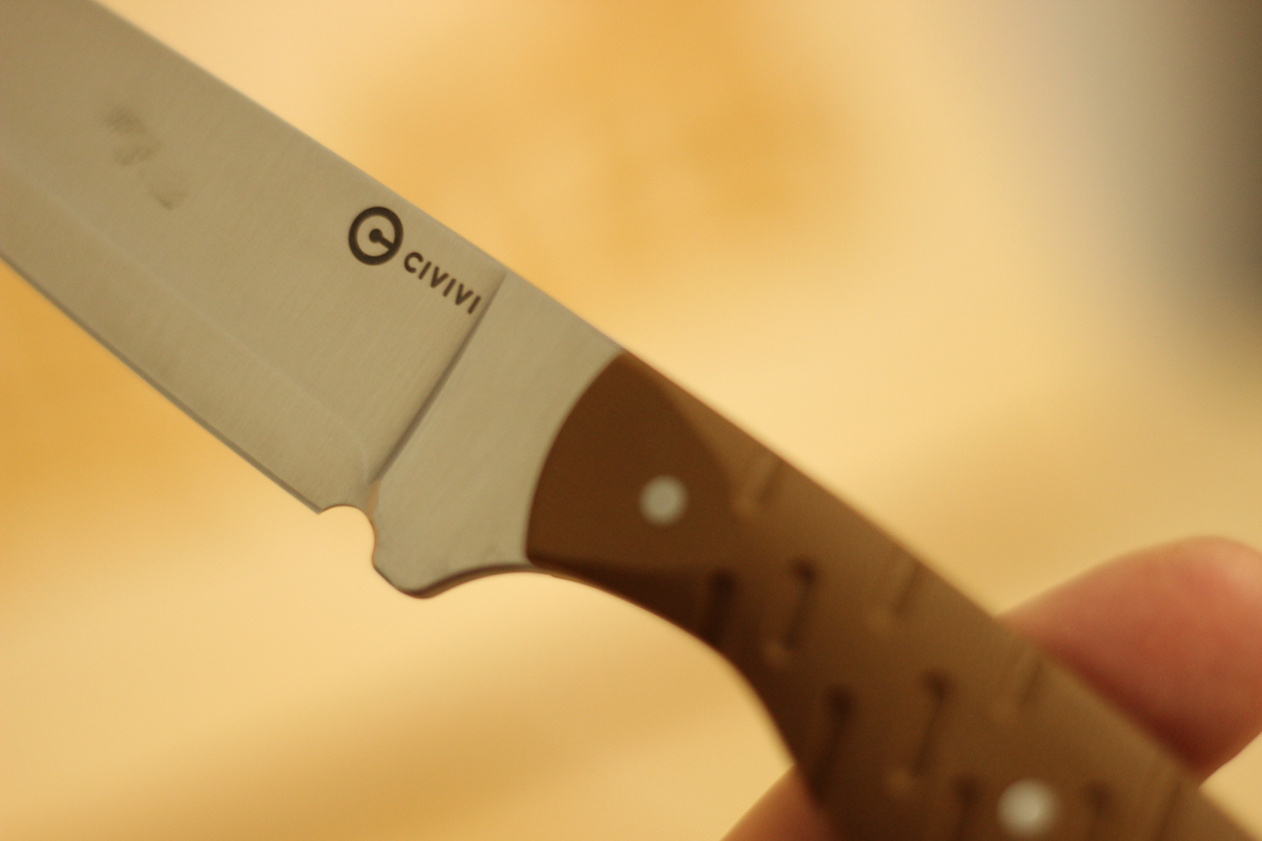 Extreme close-up of the Civivi logo etched on the satin blade flat just behind the grind transition, with the brown G10 handle and a stainless pin visible at the right edge