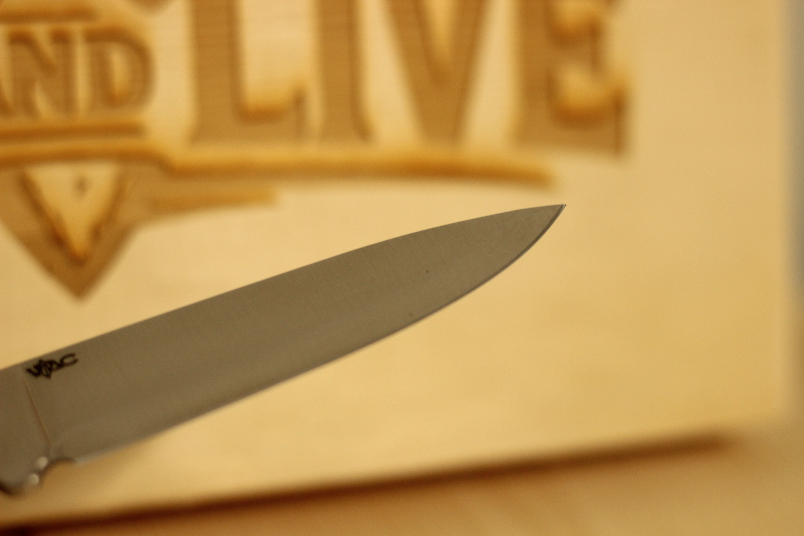 Close-up of the satin blade tip showing the small VTAC wreath-and-arrow logo etched into the steel, with the Hunt & Live wood-burned logo blurred in the background