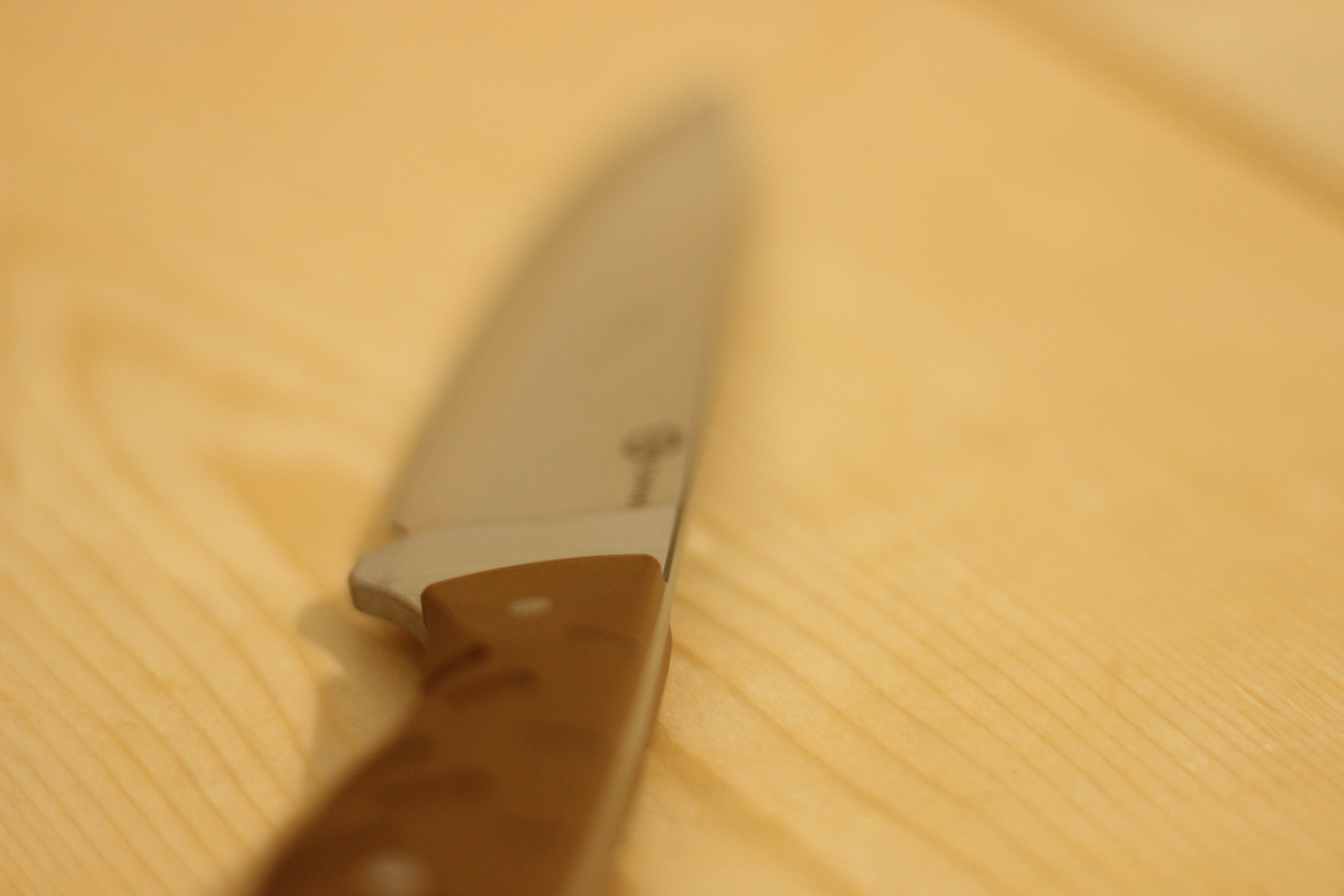 The blade laid on a wood plank surface from a high angle, the satin flat catching the light along its length, the brown G10 handle visible at the bottom of frame