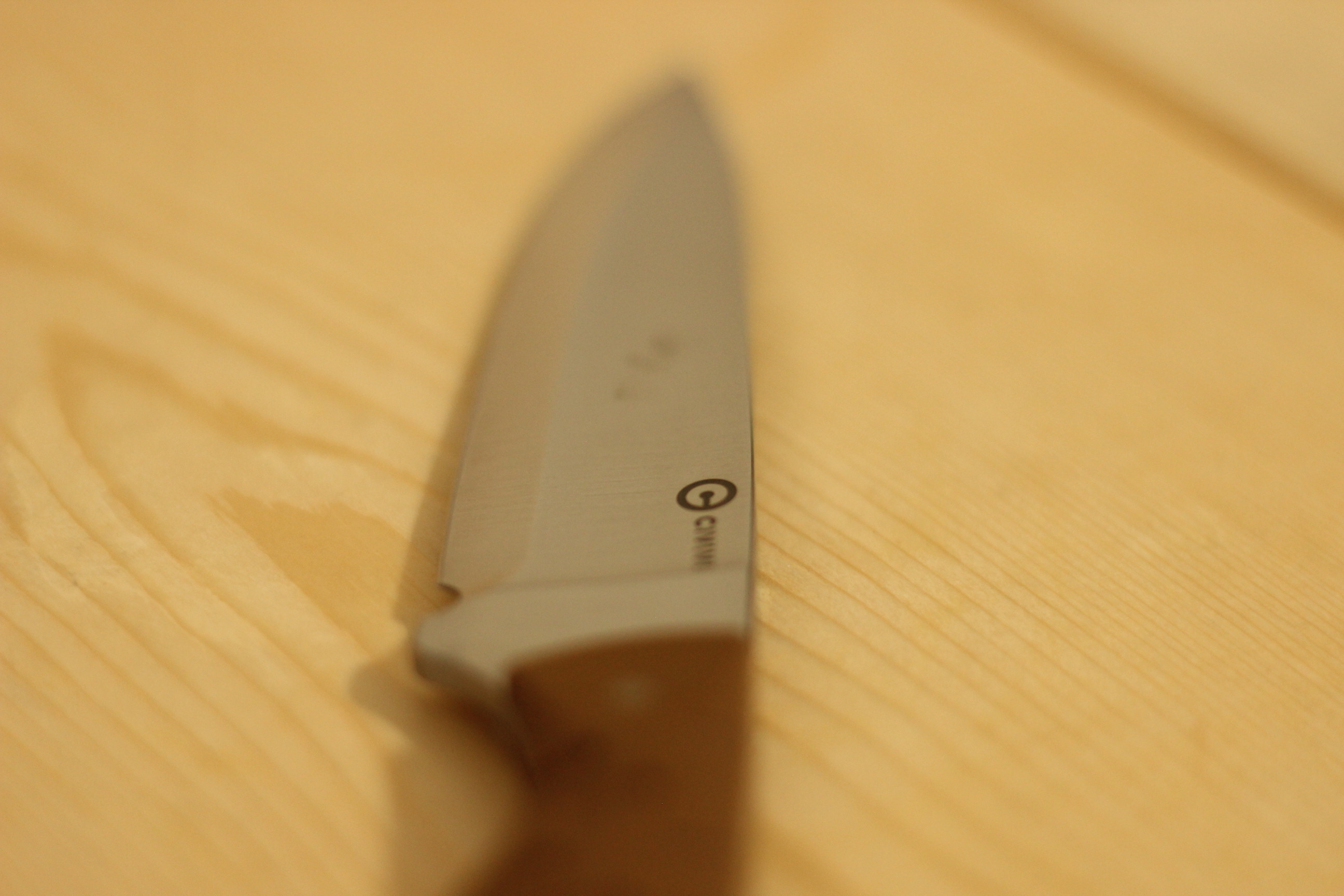 The Civivi MDRN Hunter laid on its side on a light wood plank surface showing the satin blade flat and the small Civivi C-logo mark etched on the steel