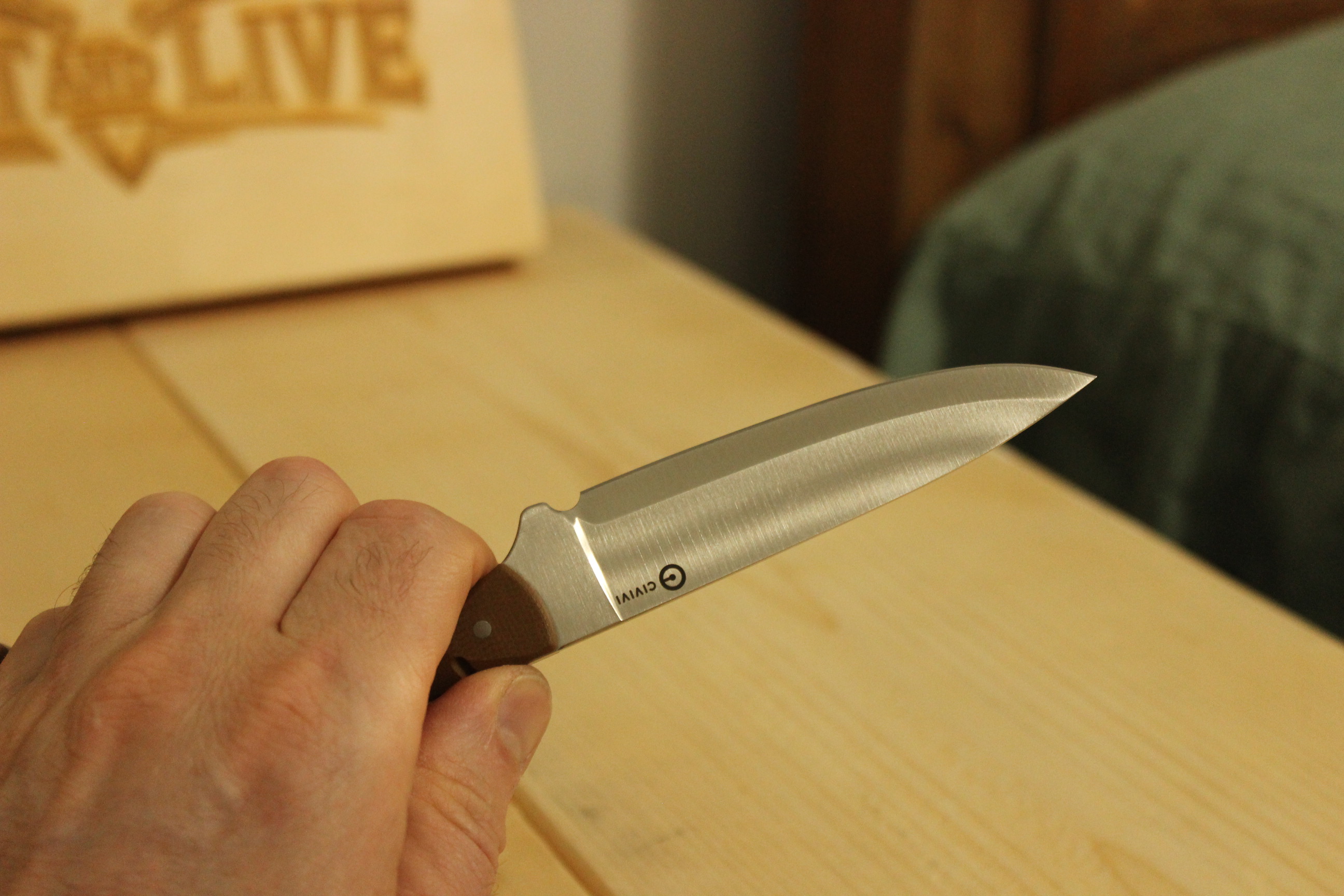 The Civivi MDRN Hunter held extended toward the camera in hand against the Hunt & Live wood-burned logo sign in the background, full blade and brown G10 handle visible in clean profile