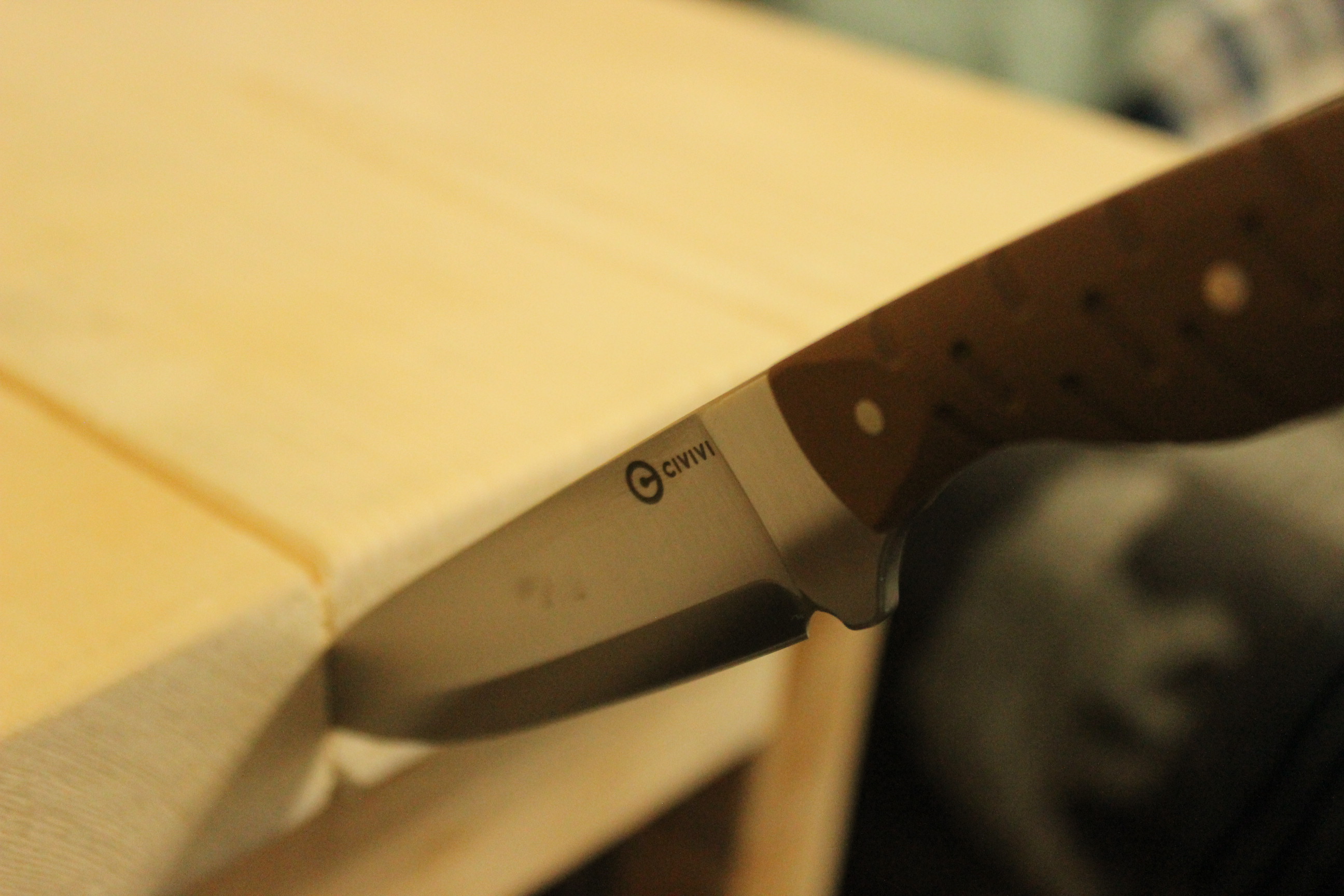 The MDRN Hunter laid edge-down against the corner of a wood workbench with the Civivi mark visible on the satin blade flat and the brown G10 handle resting against the edge