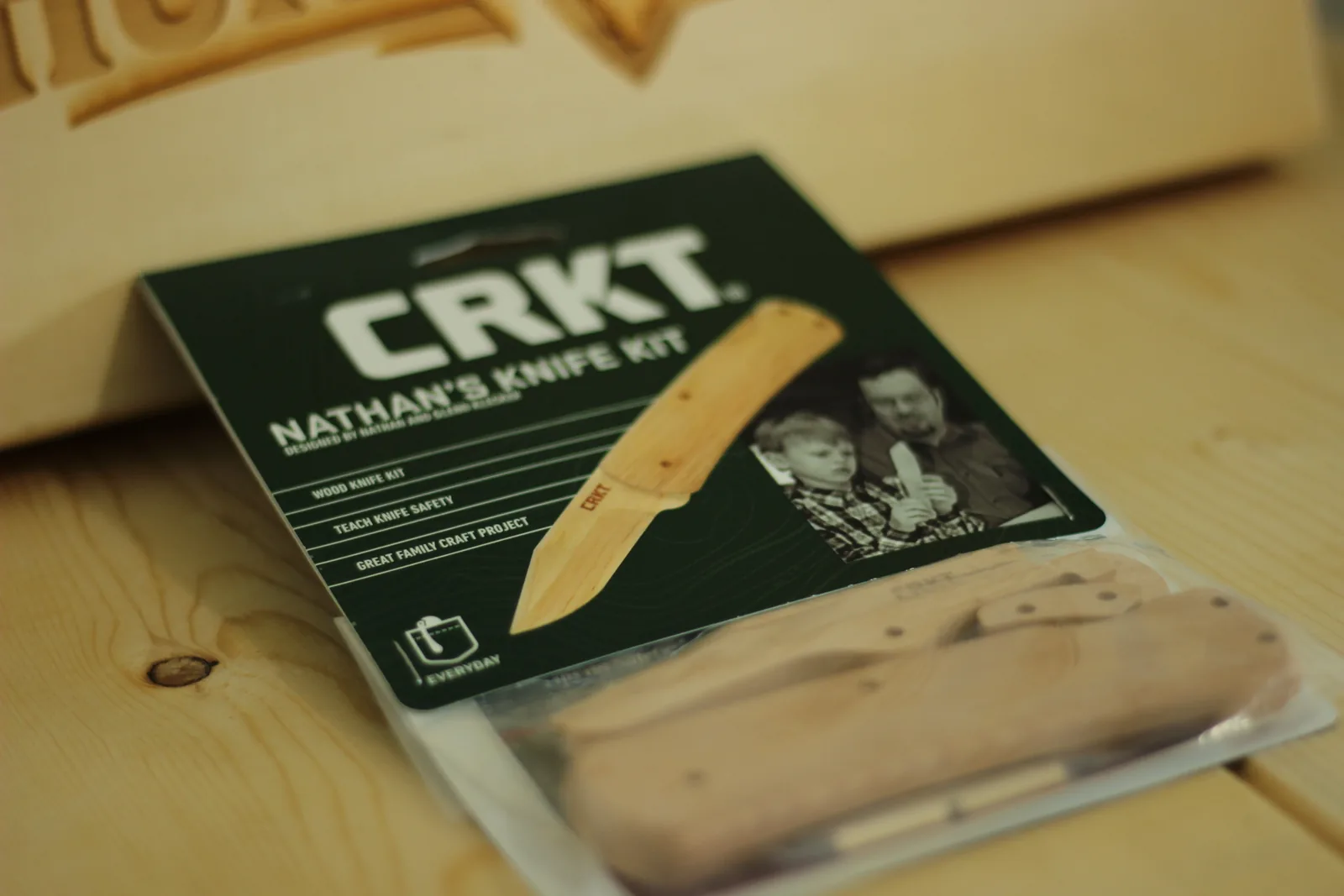 The green CRKT Nathan's Knife Kit package lying on a pale wooden bench with a laser-engraved wooden plaque behind it