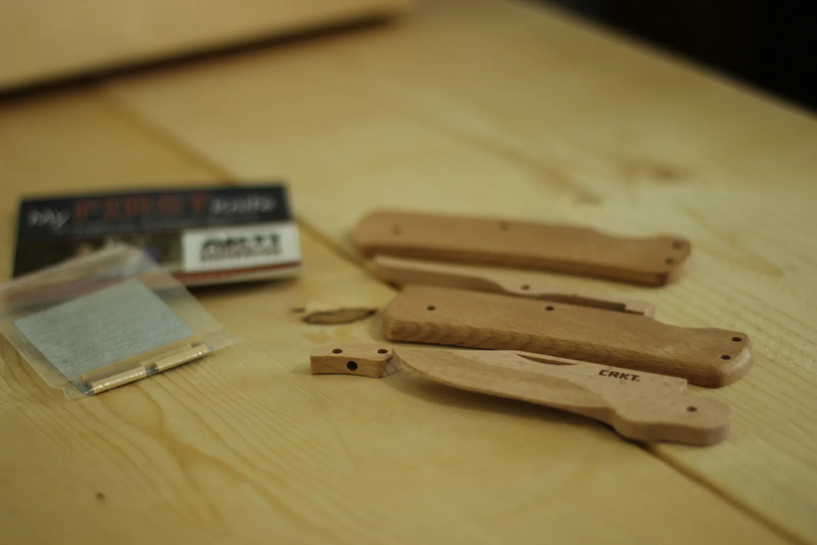 All the beech parts of the kit laid out on a wooden bench: two handle scales, the blade, the lock bar, a small spacer block, plus a bag of dowels and sandpaper and the safety booklet