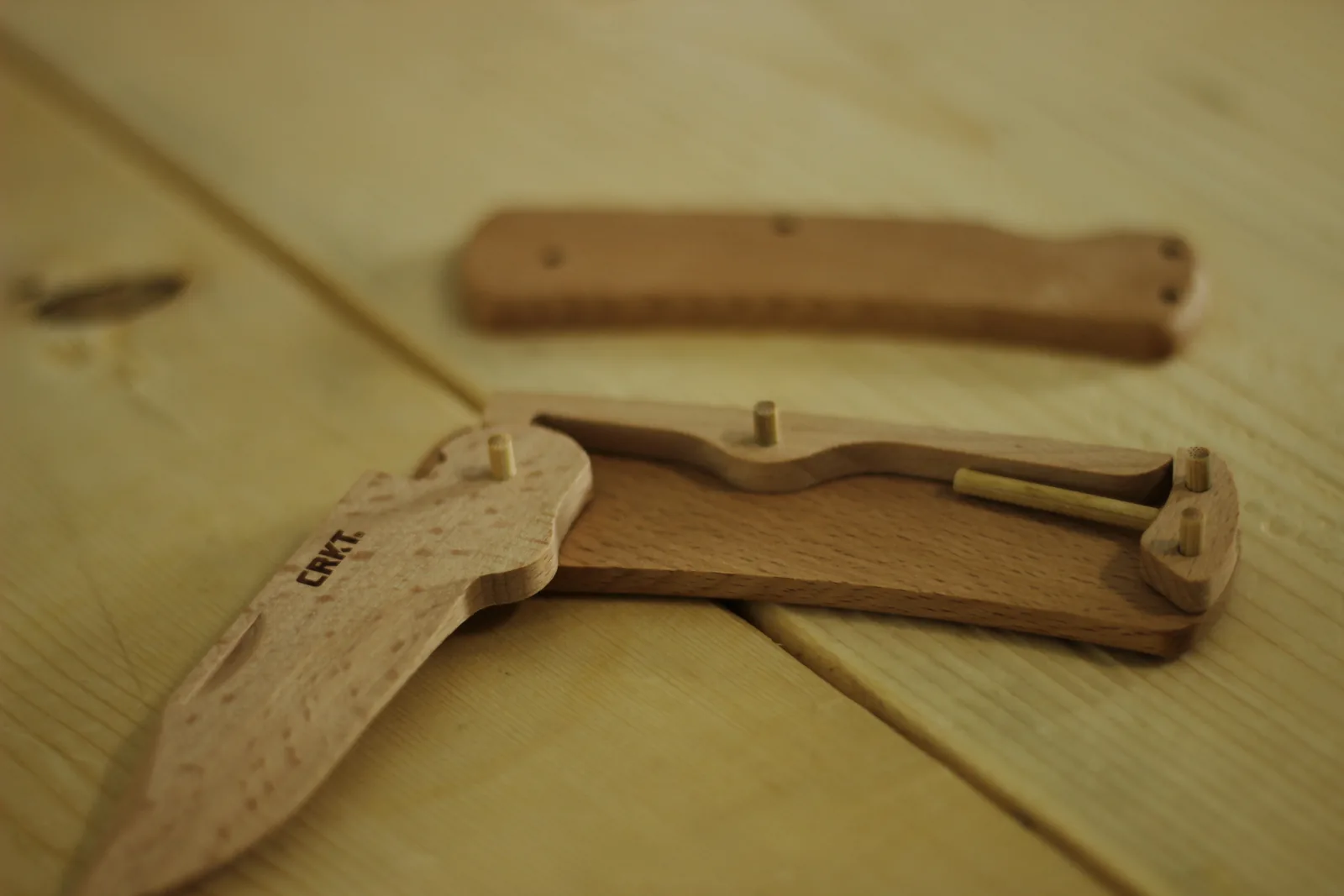 One handle scale lying flat with the shaped lock bar and a long dowel positioned inside it, pins standing upright, and the blade attached at the pivot