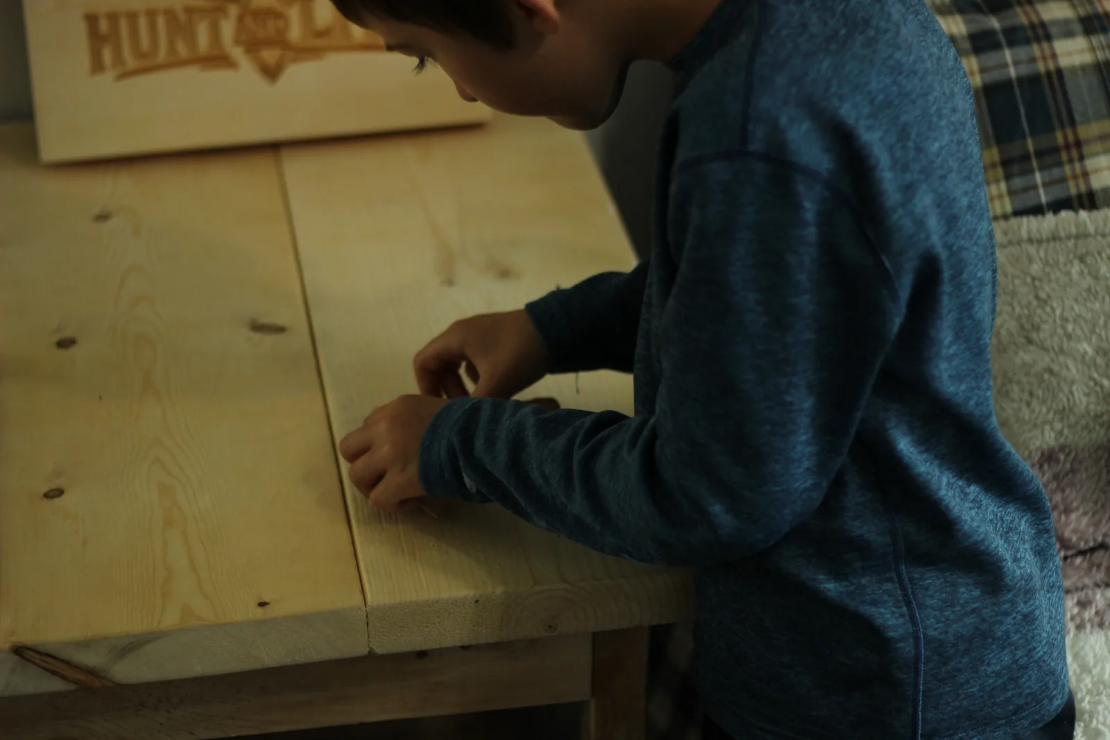 A child in a blue jumper leaning over a pale wooden workbench working on the kit, with a Hunt and Live engraved plaque propped behind