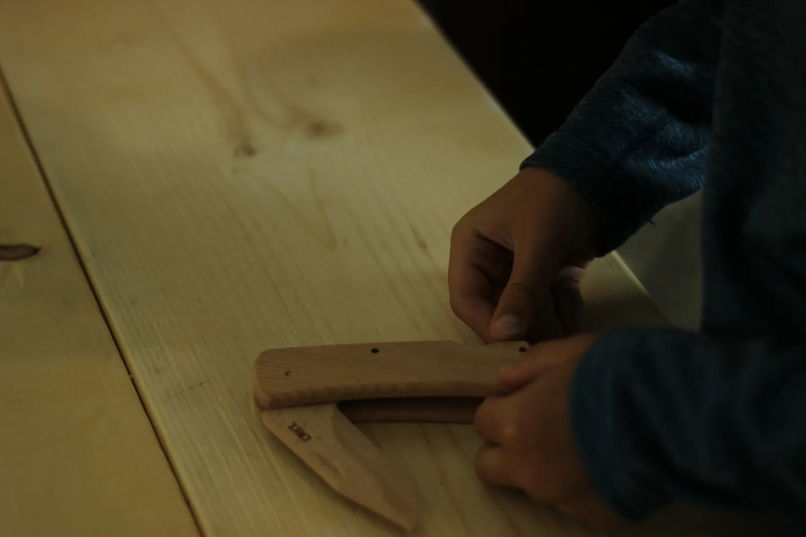 Two hands lining a beech handle scale up over the standing pins of the part-built knife on a workbench