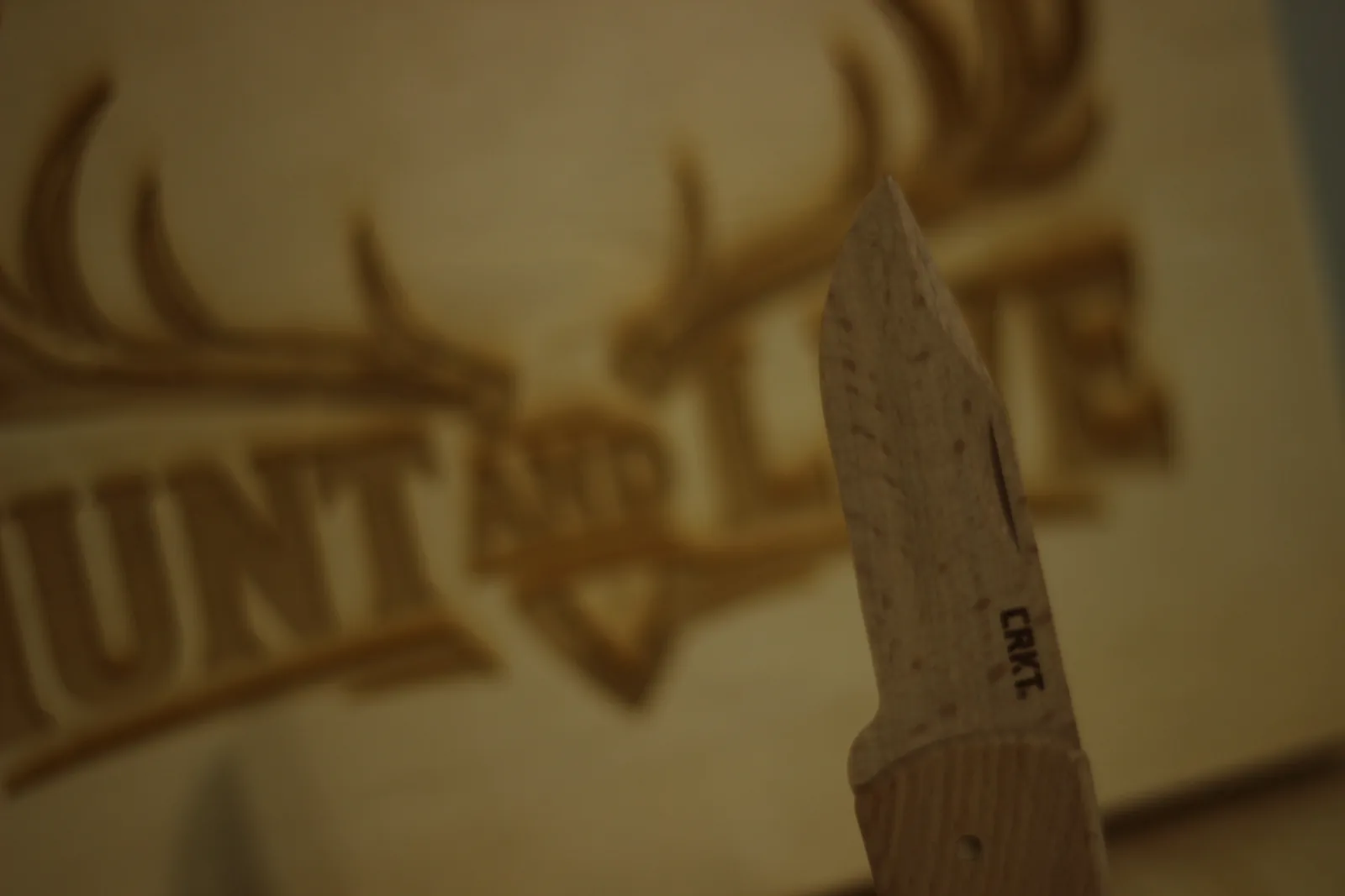 The wooden blade tip held up in front of a blurred laser-engraved Hunt and Live wooden sign