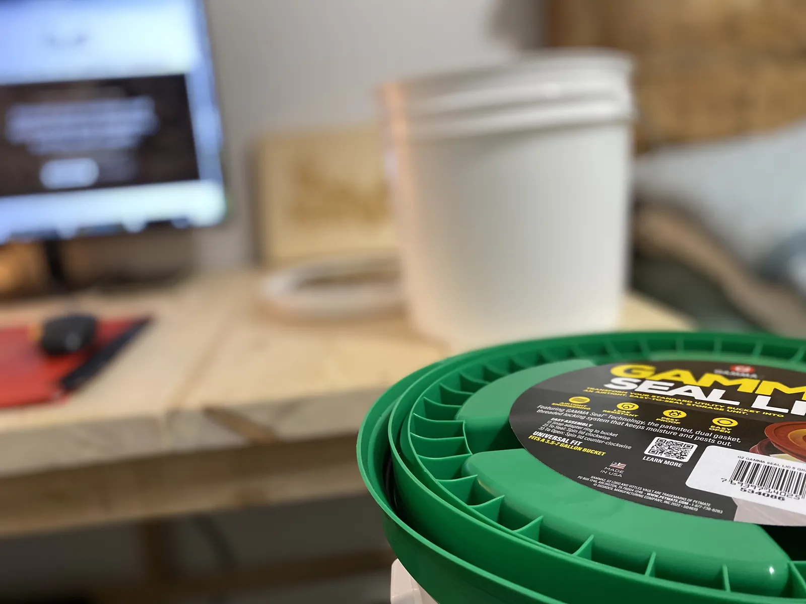GAMMA Seal Lid in green with the retail label visible, sitting on the workbench beside a computer screen, white 5 gallon bucket and Hunt & Live plaque