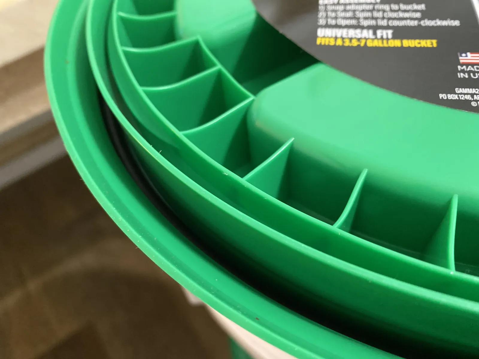 Diagonal close-up of the GAMMA Seal Lid showing the lid teeth, dual-gasket profile, "UNIVERSAL FIT FITS A 3.5-7 GALLON BUCKET" and the small "MADE IN USA" flag