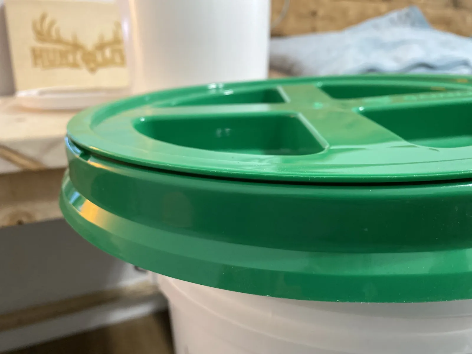 GAMMA Seal Lid resting on top of a white bucket showing the "CLOSE" arrow with the Hunt & Live plaque visible in the soft background