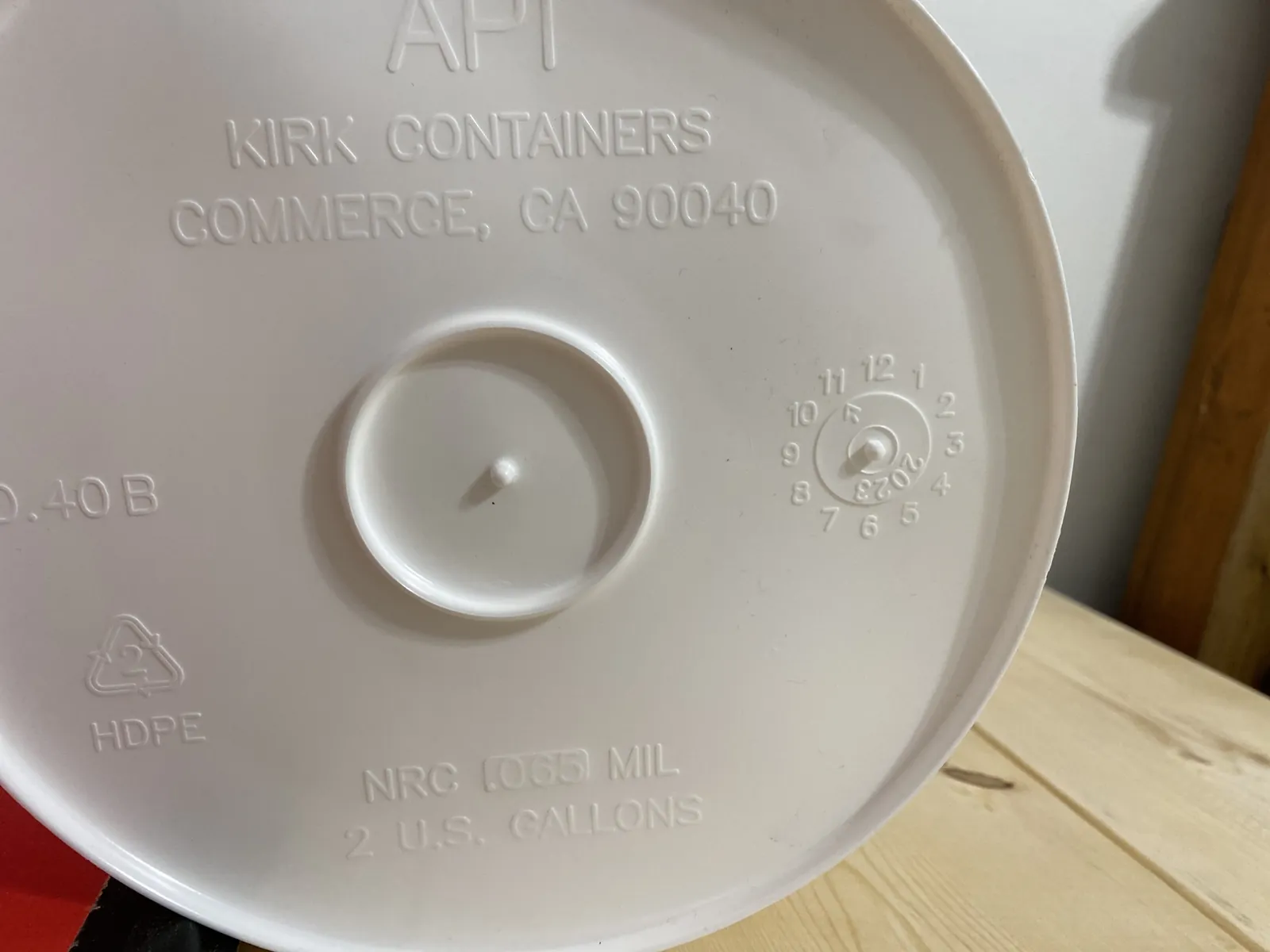 Close-up of a white 5-gallon food-grade bucket rim showing the heavy rolled-edge profile and the molded GAMMA-compatible rim ridges with a partial WARNING label visible at the base