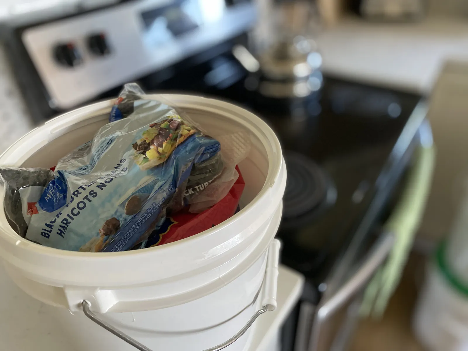 A white 5-gallon bucket on a black stovetop in a homestead kitchen, holding bags of MERIT Bleu black turtle beans / haricots noirs and other dry-goods packets ready to be sealed under the GAMMA Seal Lid