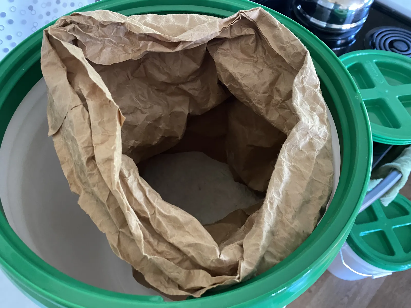Three white food-grade buckets stacked vertically in a kitchen, the lower two sealed with green GAMMA Seal Lids and the top bucket without one — sitting beside a stainless gas/electric range