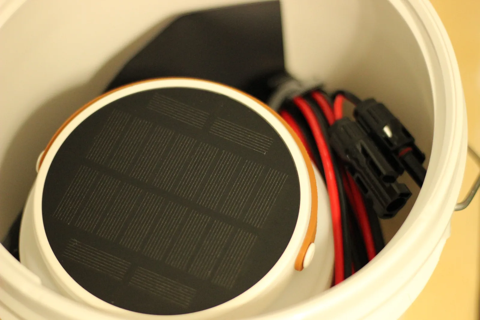 Inside view of a white bucket holding a folded portable monocrystalline solar panel with orange edge band, MC4 connectors, and red-and-black PV wiring — sealed dry storage for off-grid solar gear