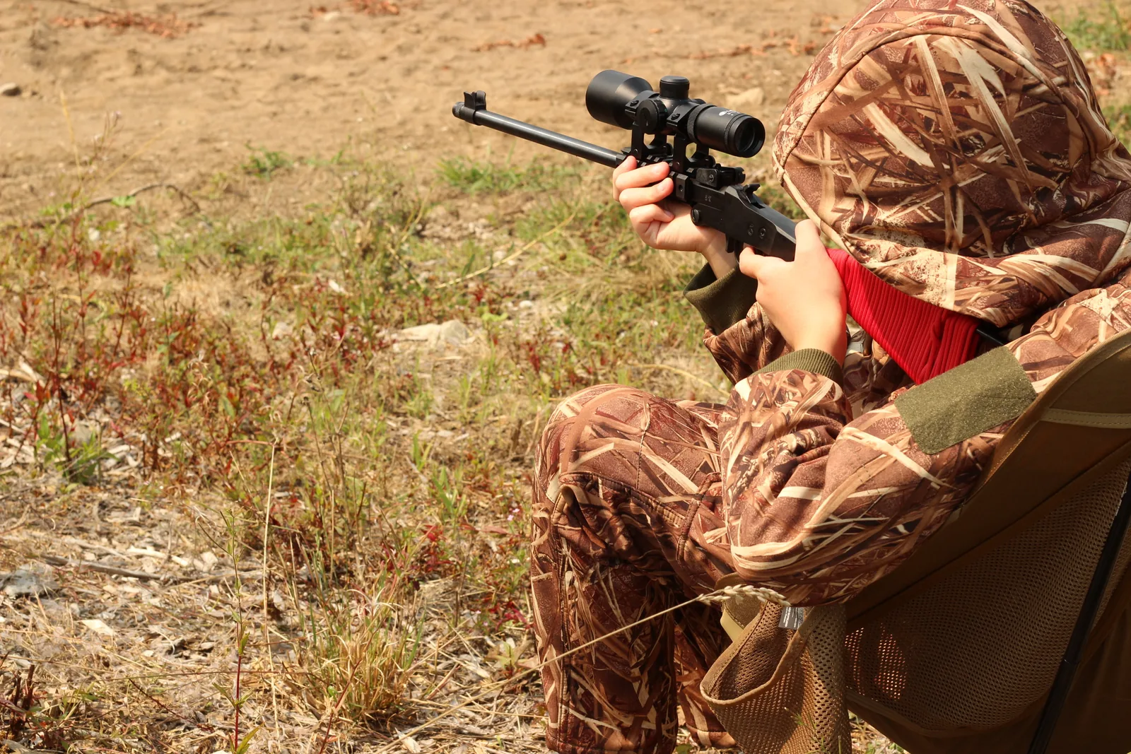 A shooter in a leafy ghillie suit seated in the coyote-brown Helikon-Tex Traveler chair on a dry hillside, scoped rifle shouldered and pointed downslope