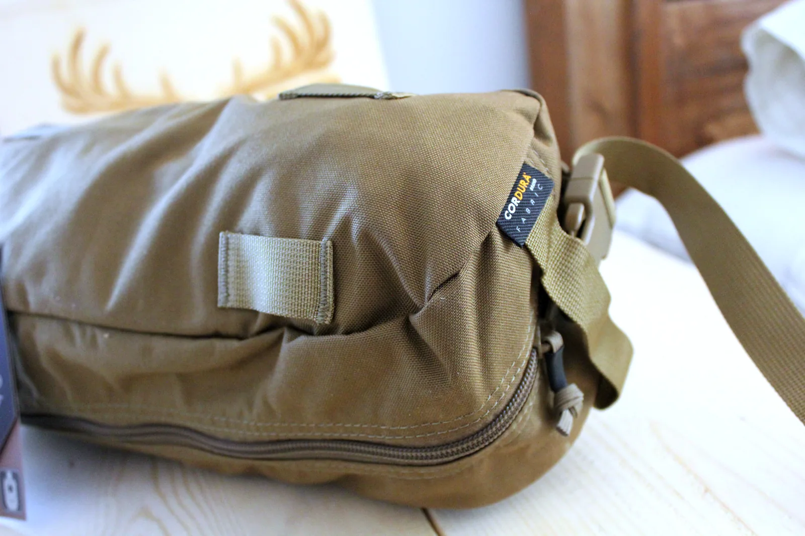 The olive-green Cordura carry bag zipped shut on a pale wooden bench, a webbing shoulder strap running off the end
