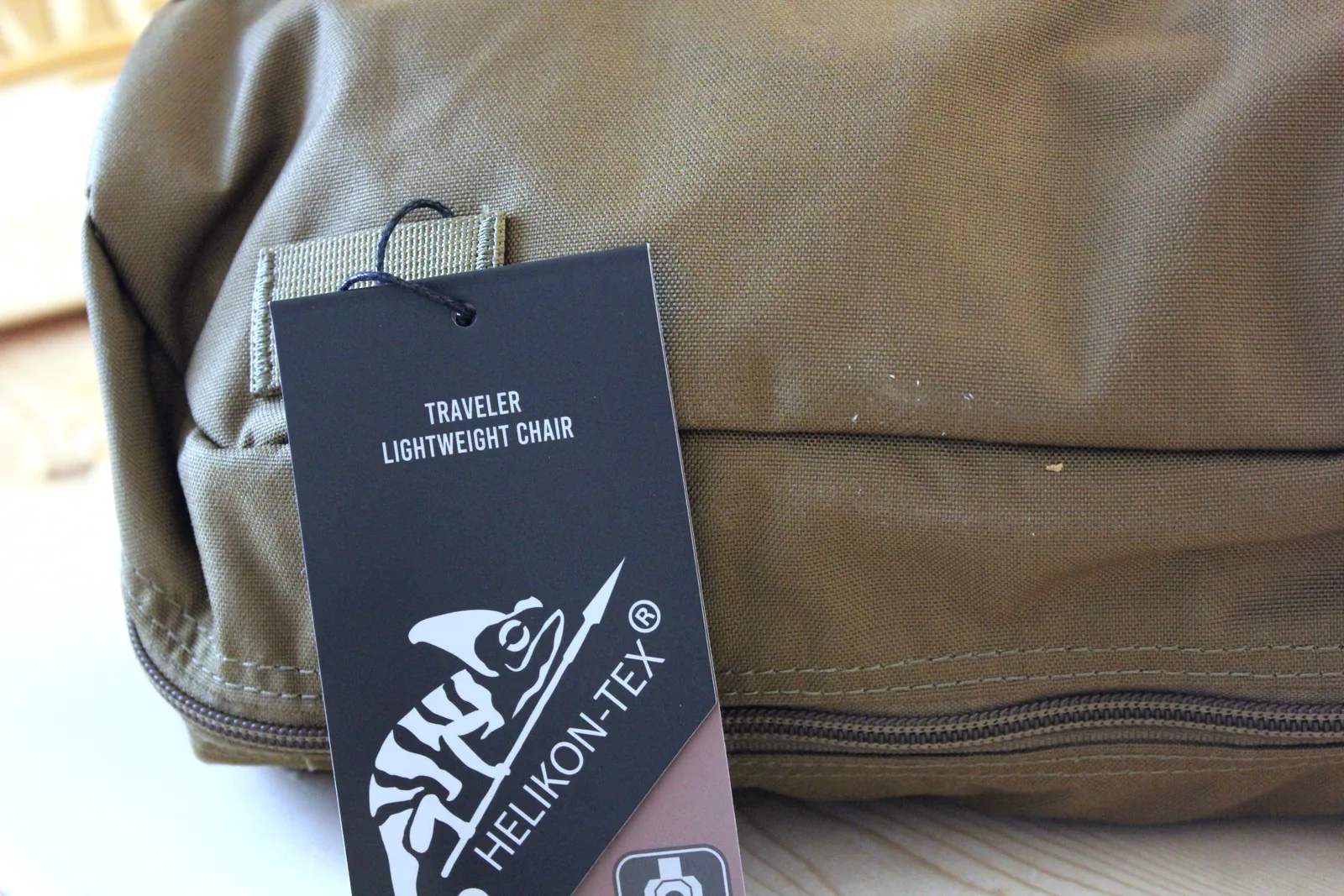 A black Helikon-Tex hang tag reading TRAVELER LIGHTWEIGHT CHAIR attached to the coyote carry bag