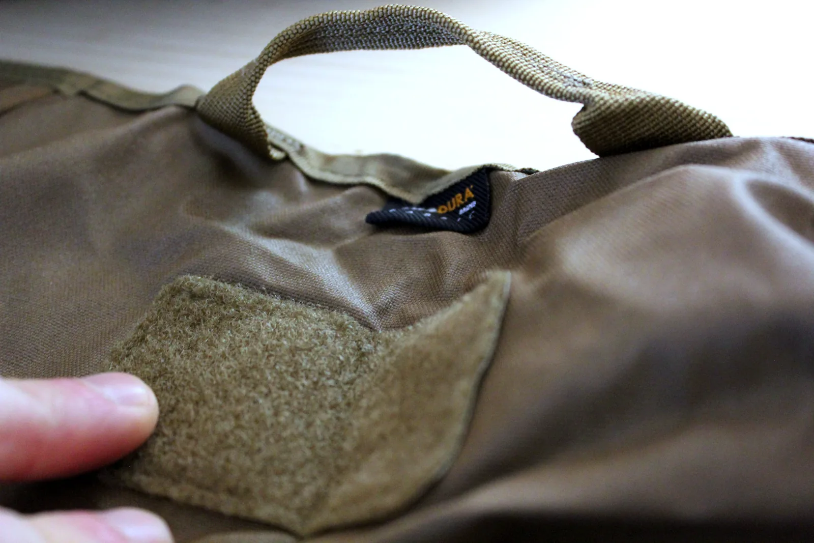 A hand lifting the corner of a square hook-and-loop patch panel on the back of the seat, with a webbing haul loop above it