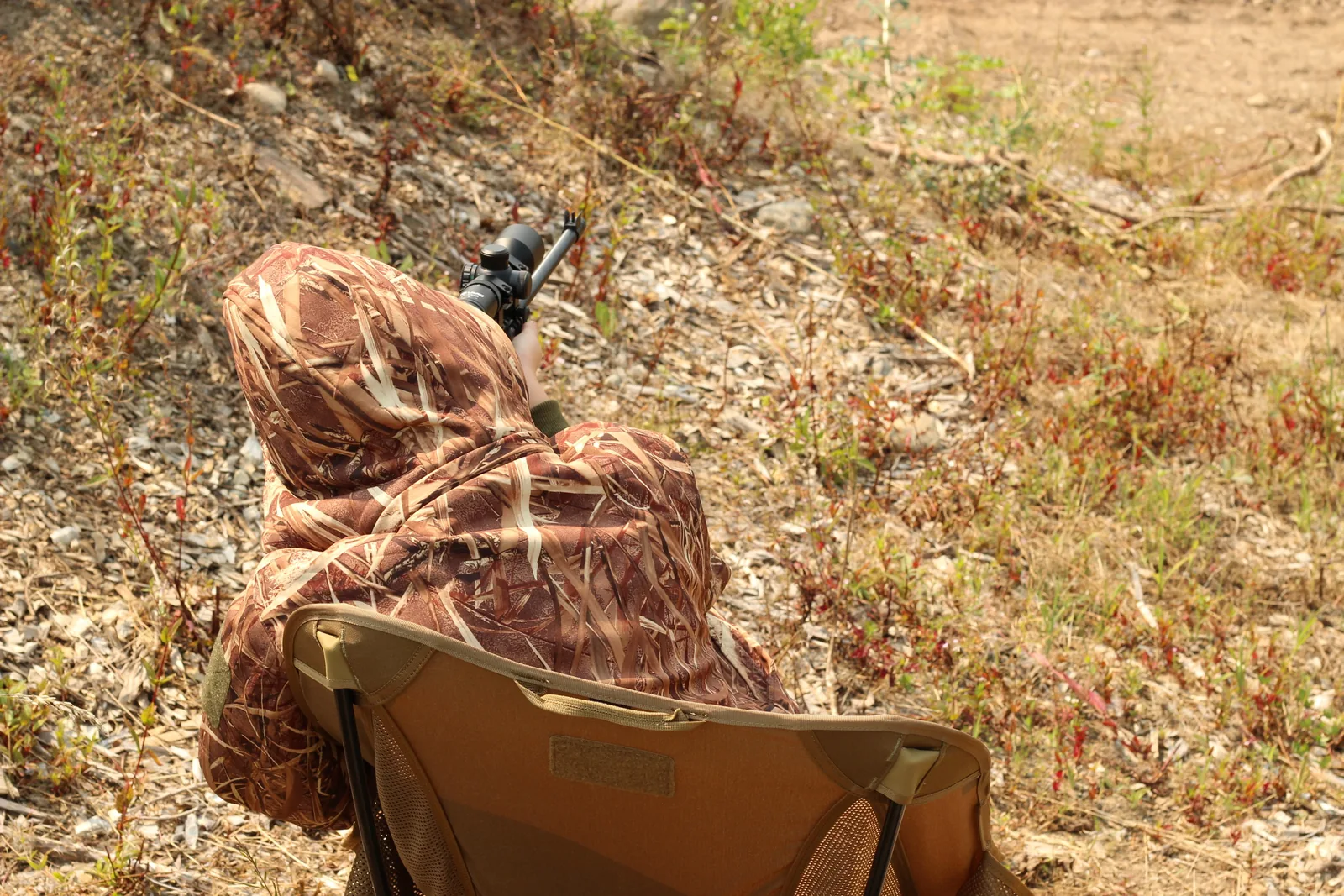 Side view of a seated shooter in a leafy suit with a scoped rifle up, the coyote chair frame visible beneath