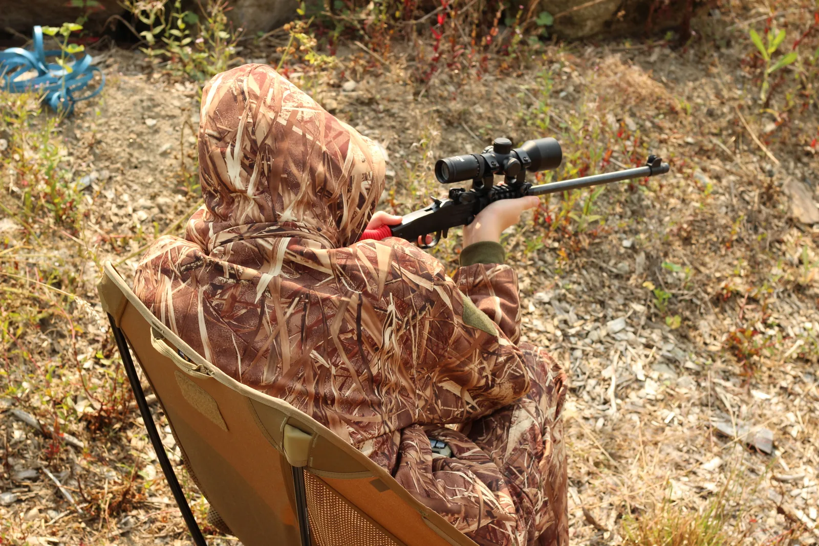 Over-the-shoulder view of a seated shooter in the chair with a scoped rifle pointed downslope across dry ground