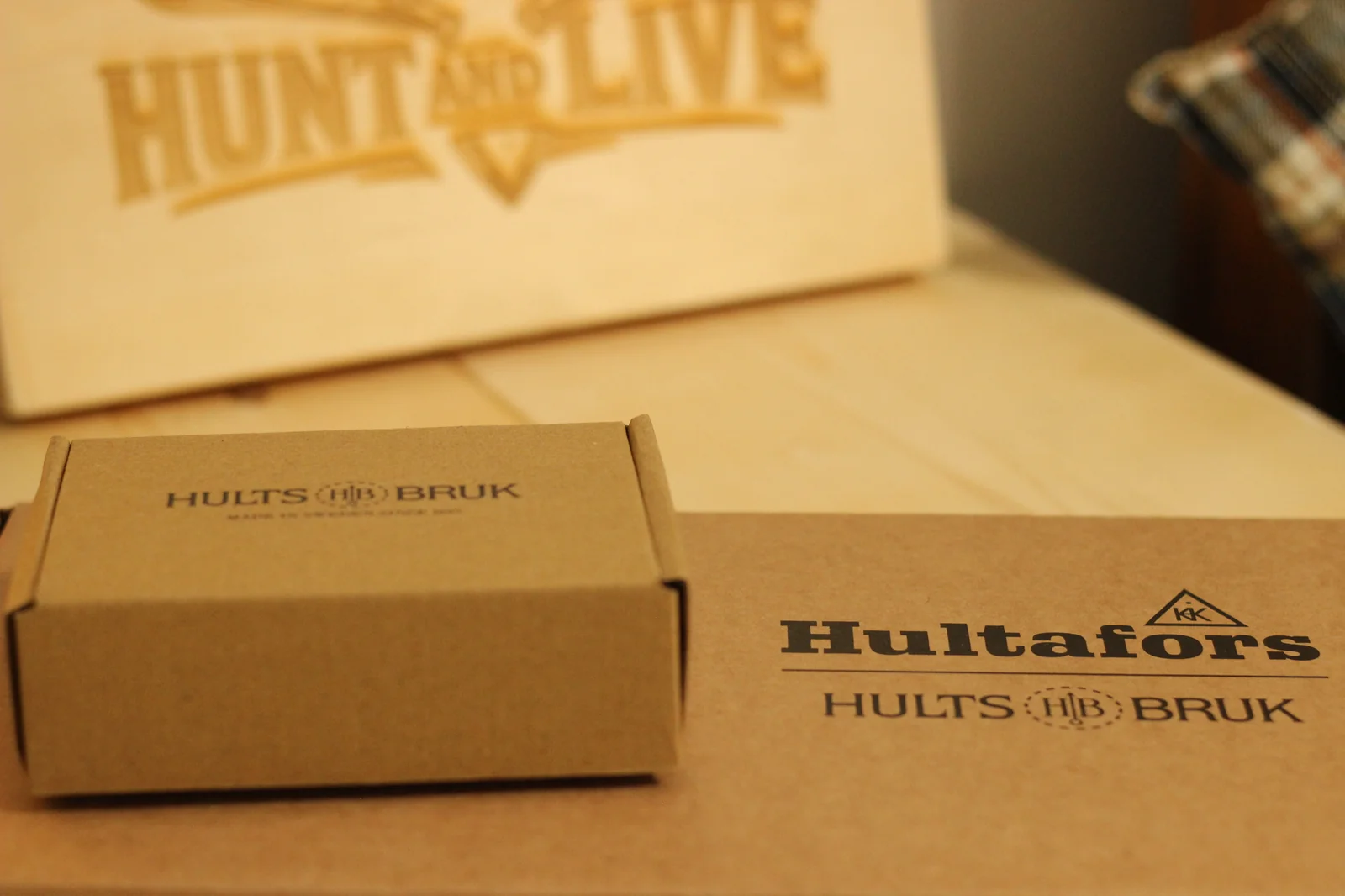 Small Hults Bruk grinding stone box and larger Hultafors Premium Axes box on a wooden surface with the Hunt and Live wooden plaque behind