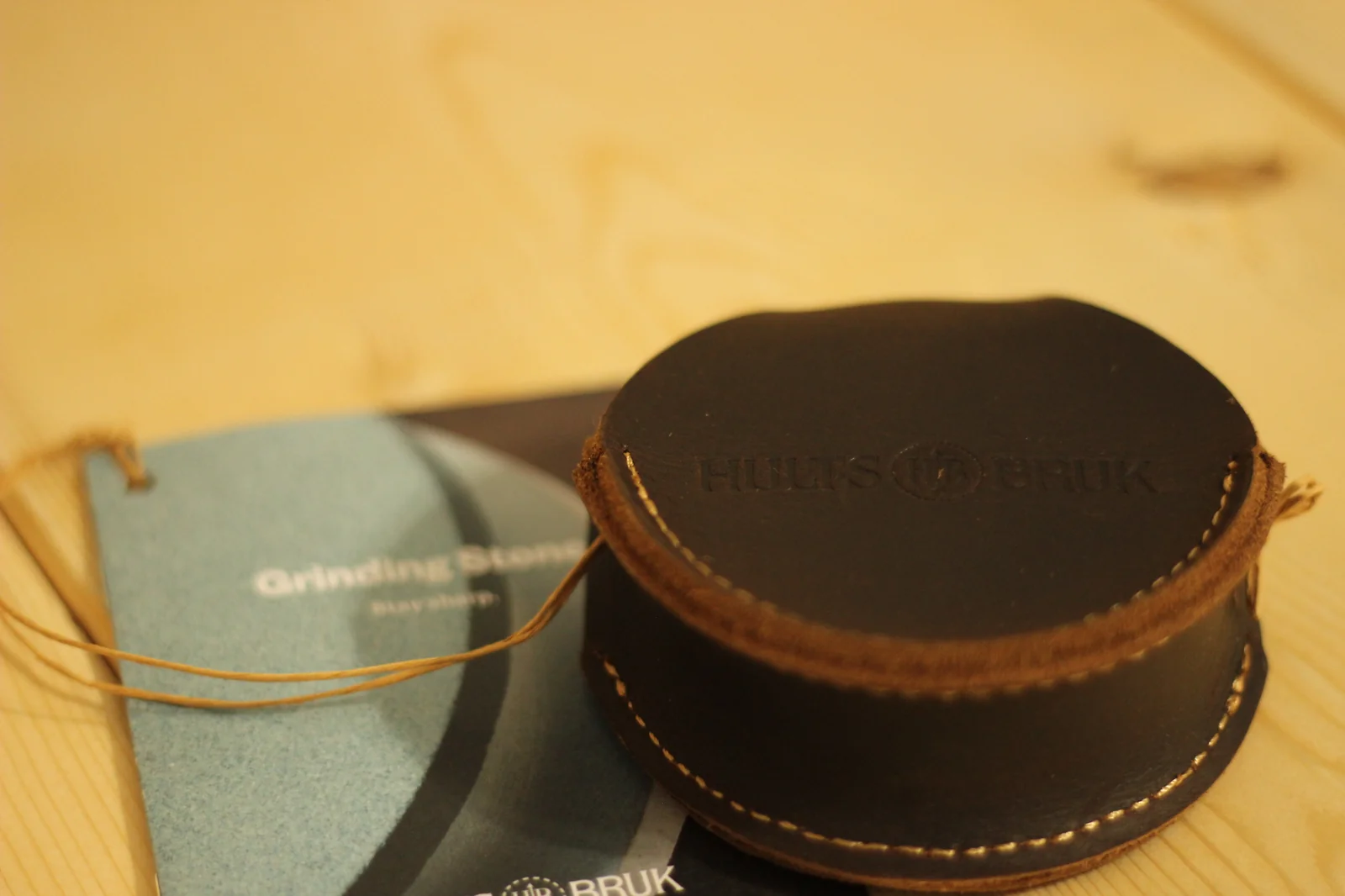 Round leather Hults Bruk grinding stone pouch sitting on a black product card showing the stone, with the HB embossed logo visible on the leather