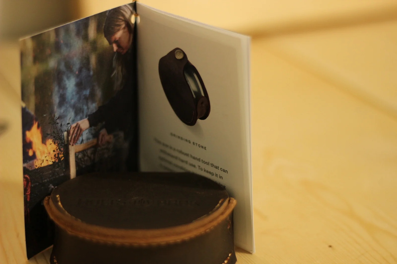 Open booklet propped above the grinding stone pouch showing a photograph of a person sharpening at a campfire on the left page and a product shot of the leather pouch on the right