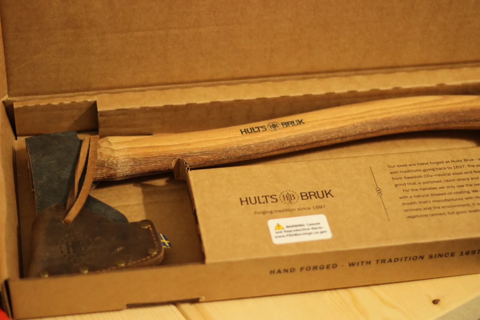 Open Premium Axes box showing the Dvärdala axe inside with leather sheath on the head and product booklet visible alongside, with the HAND FORGED WITH TRADITION SINCE 1697 text printed on the inside box edge