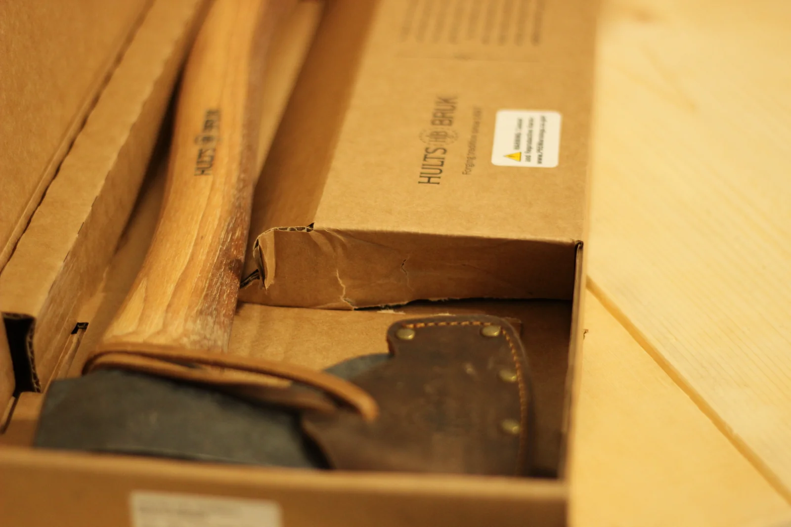 View along the axe inside the box showing the hickory handle with the HULTS BRUK brand mark and a yellow warning sticker on the box edge