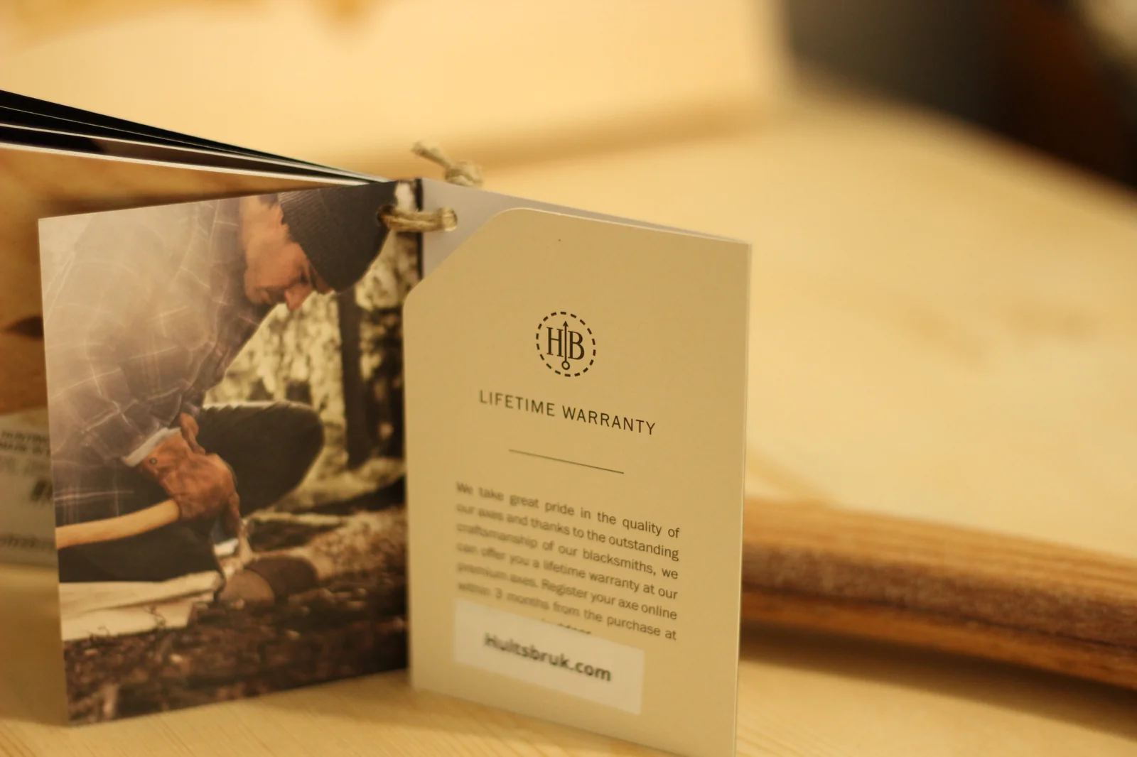 Hang tag reading LIFETIME WARRANTY with the HB hallmark and instructions to register the axe at hultsbruk.com, alongside a photo card showing a man kneeling with an axe in a snowy forest