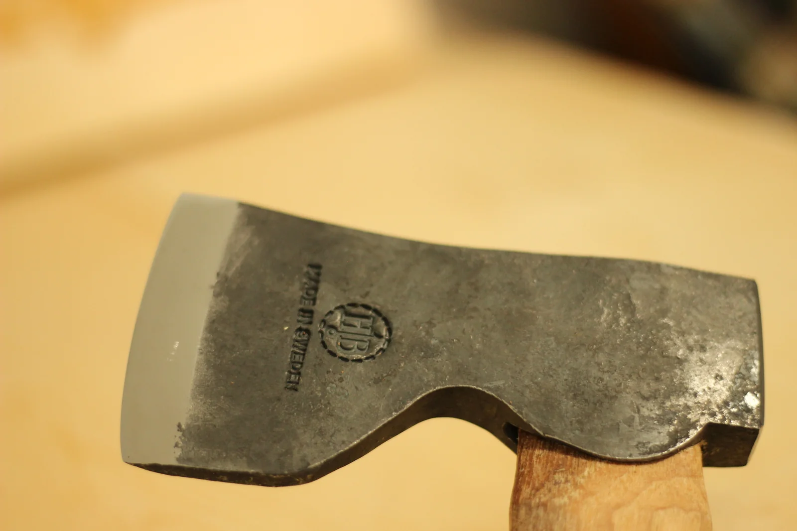Full side profile of the axe head against a pale wood background showing the blackened forged face the polished cutting edge and the HB hallmark stamp