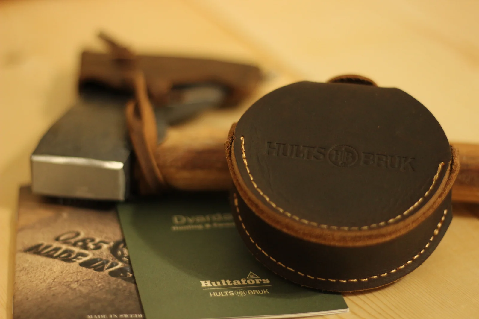 Hults Bruk leather grinding stone pouch in foreground with the axe sheath, axe head and handle, and the Dvärdala booklet arranged behind it on a wood surface