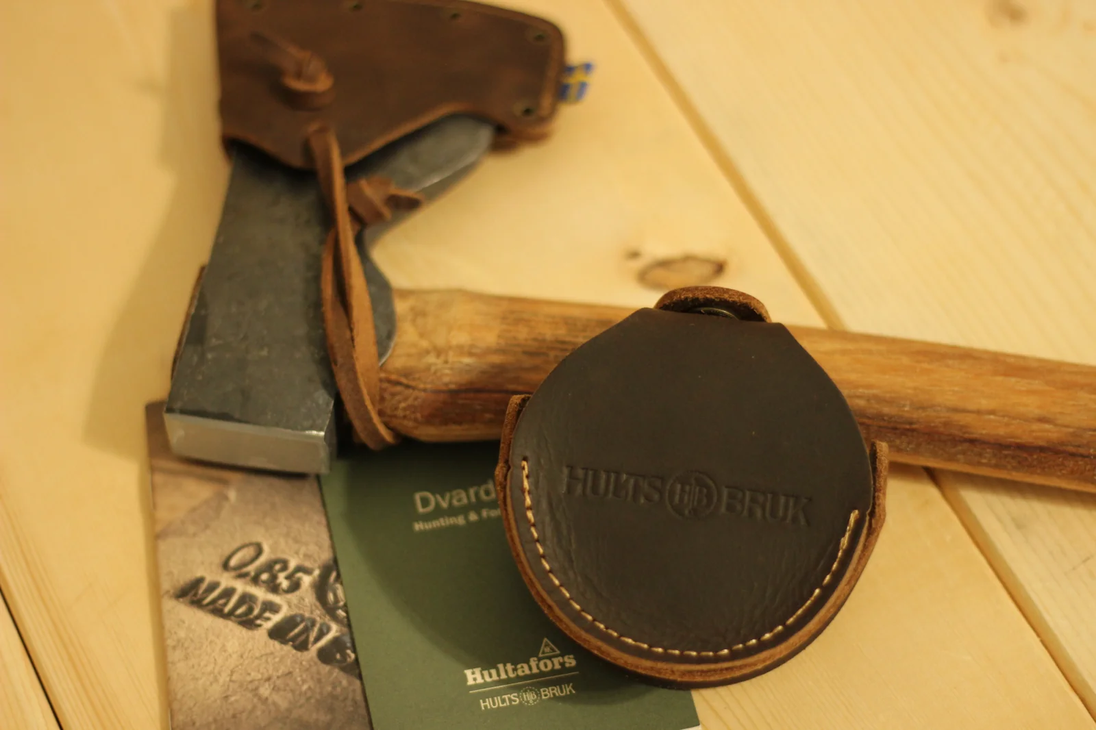 Same paired arrangement from a higher angle showing the leather grinding stone pouch the axe head in its sheath the handle and the Hultafors Hults Bruk booklet