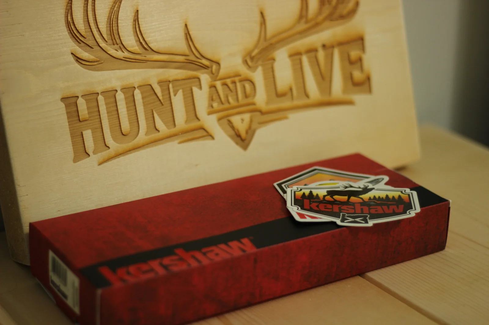 Kershaw Deschutes Combo red retail box on a wooden surface beside the Hunt and Live laser-engraved wooden plaque with antlers