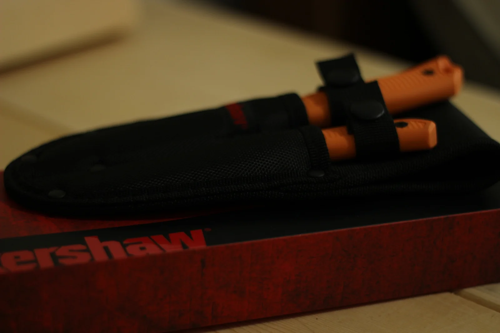 Side view of the black nylon double sheath holding both Kershaw knives with their blaze-orange handles protruding, resting on top of the red Kershaw retail box