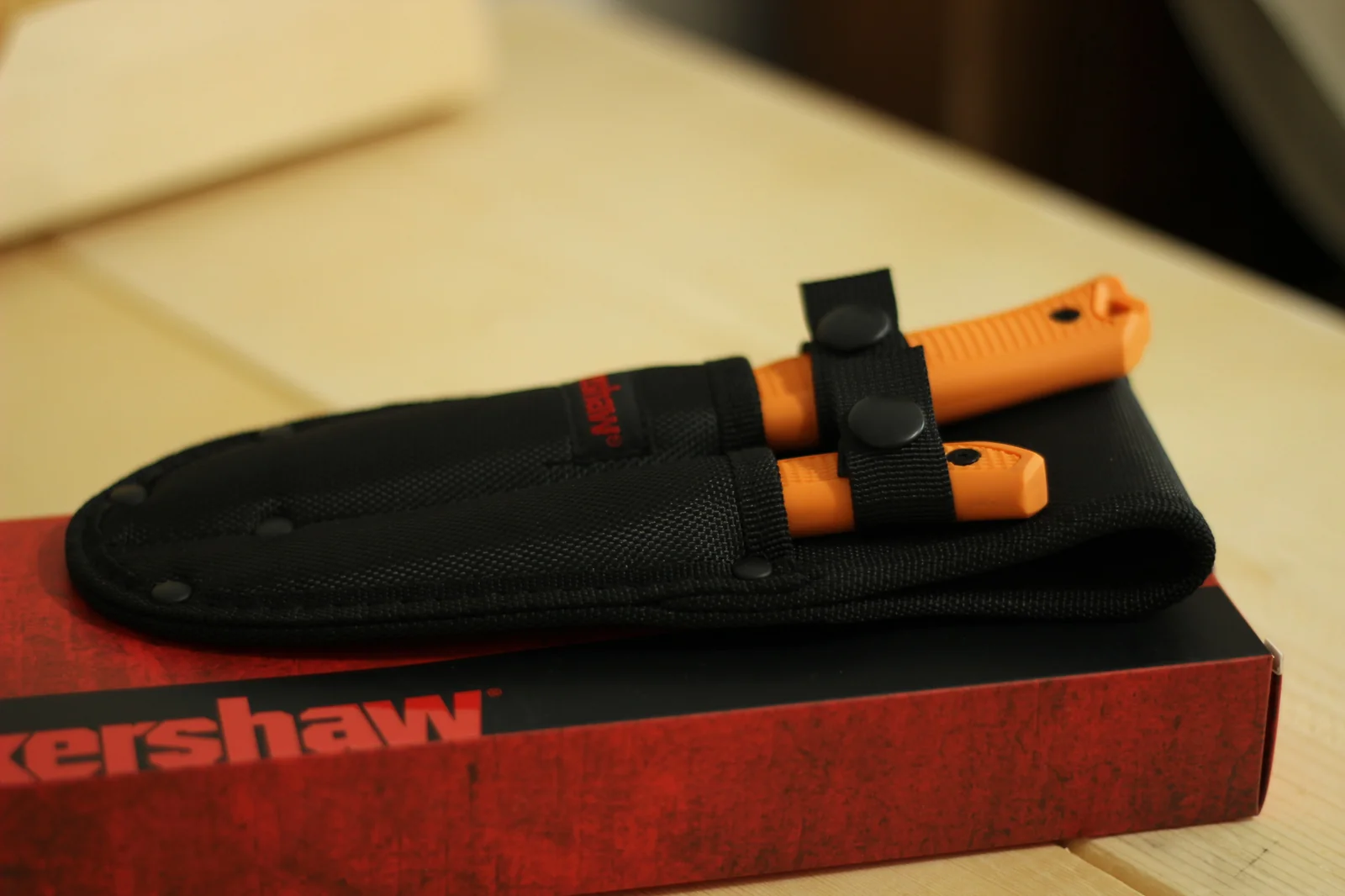 Same Kershaw double sheath holding both knives shown from a slightly higher angle on top of the red Kershaw retail box