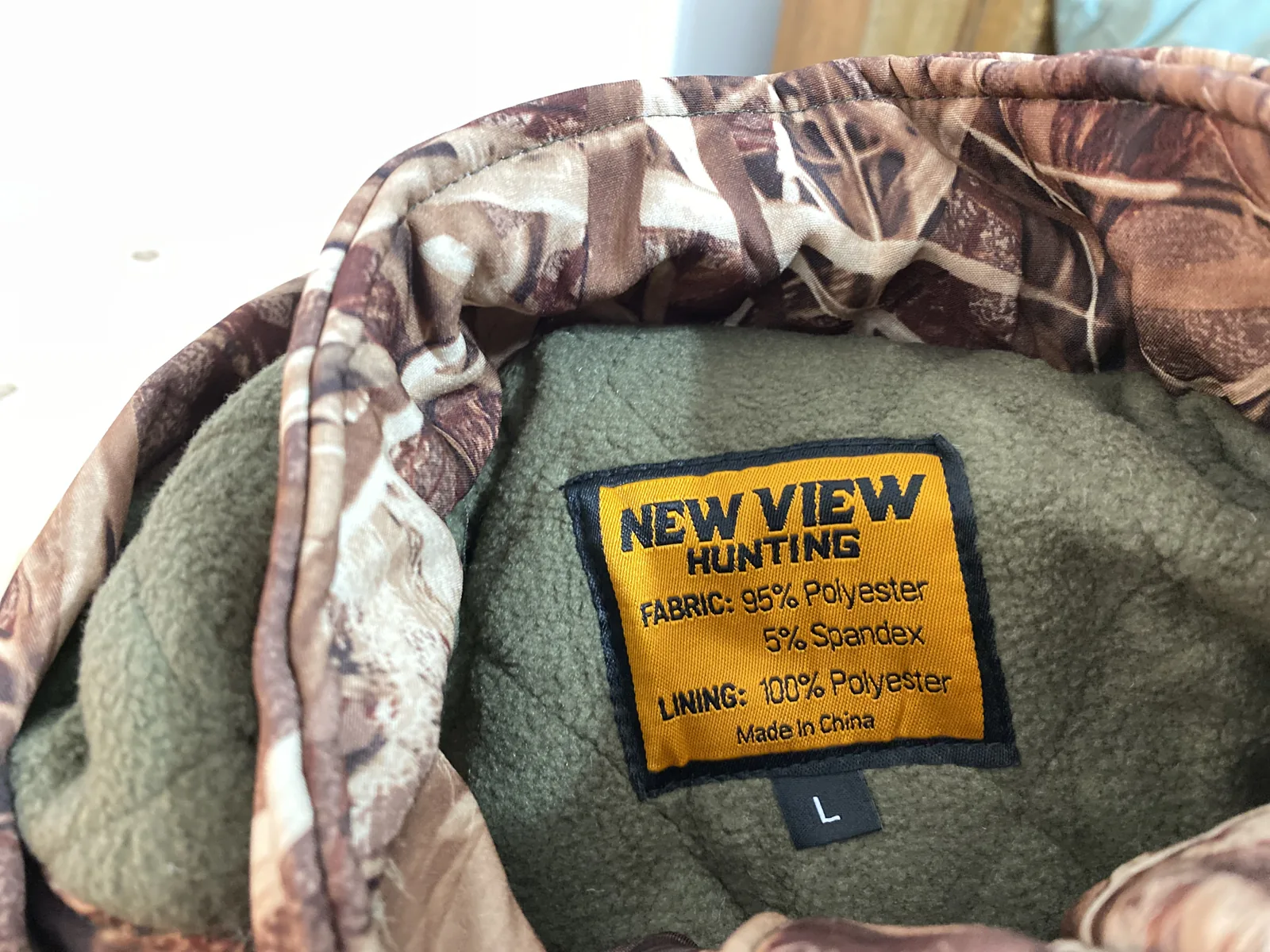 An orange and black New View Hunting neck label reading FABRIC 95% Polyester 5% Spandex, LINING 100% Polyester, Made In China, with a size L tab beneath