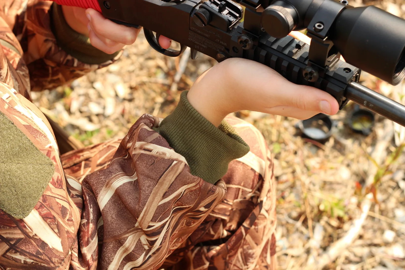 A young hunter's hands on a scoped rifle, the olive knit cuff of the camo jacket sitting neatly at the wrist