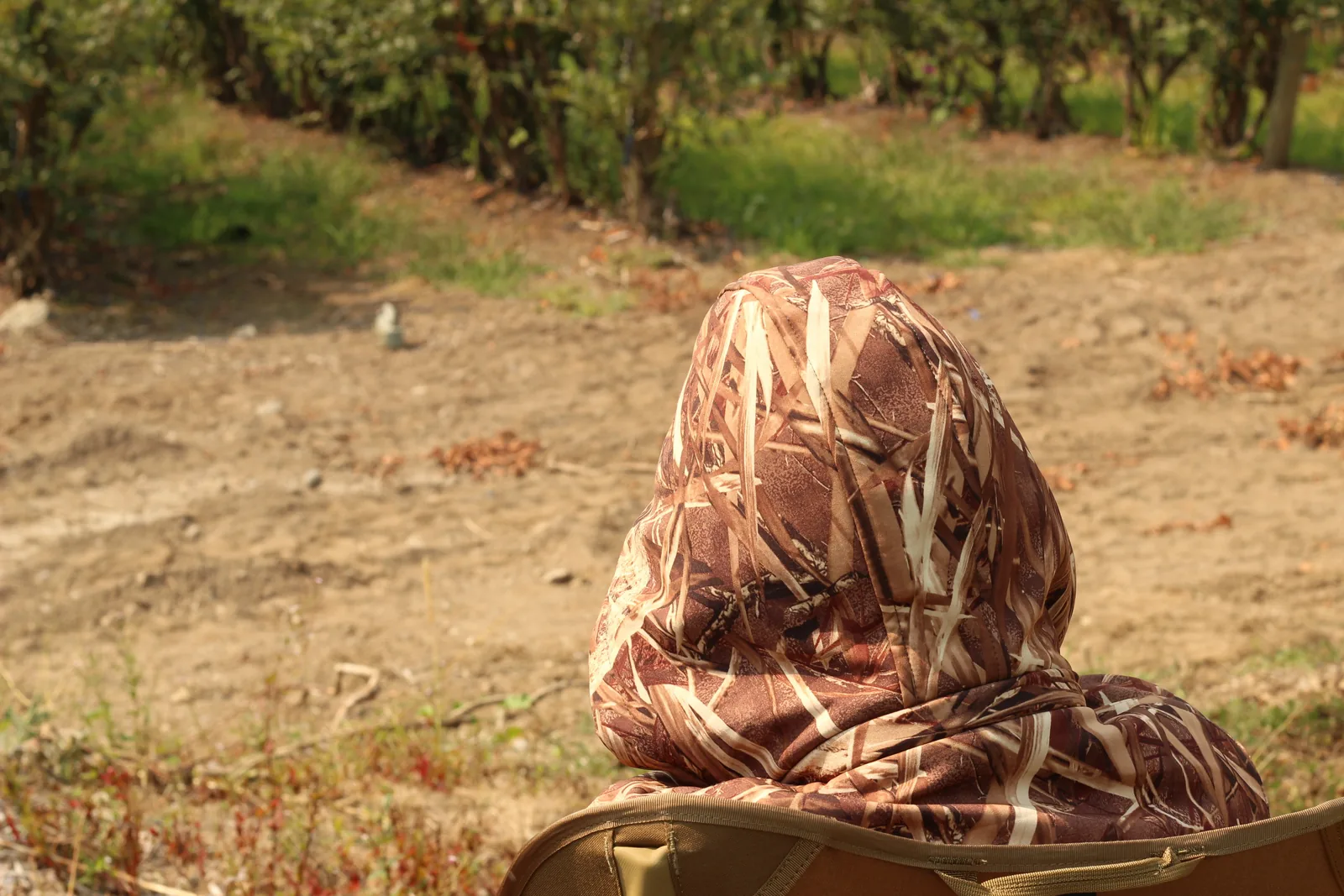 The seated young hunter from directly behind, hood up, against dry dirt and sparse grass