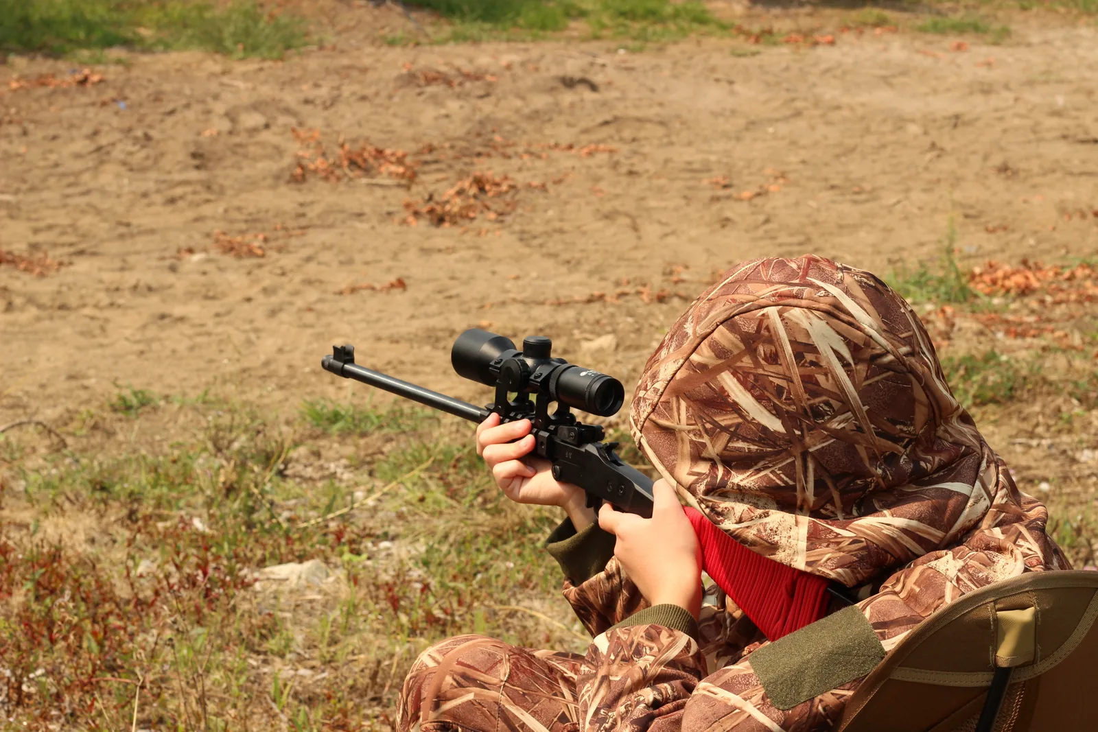 A young hunter with the scoped rifle shouldered from a seated position, hood up, in the camo suit