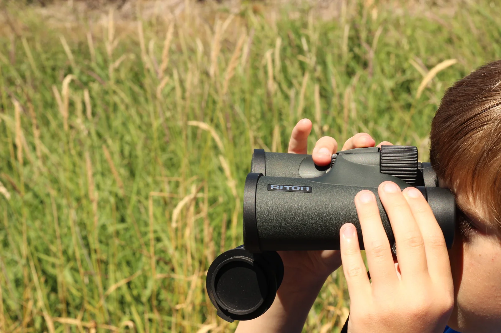 The Riton 5 Primal 10x42 ED held up to the eyes of the eleven-year-old field tester in tall summer grass, hands wrapped around both barrels with the RITON wordmark visible on the right barrel, hair just visible at the right edge of the frame, objective lens cap hanging from the strap lug